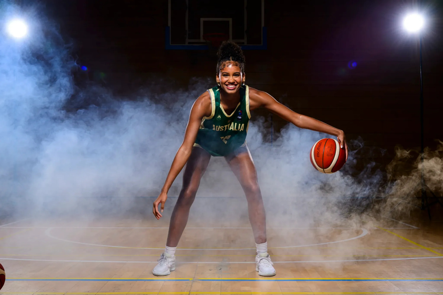 Sitaya Fagan in a green Australia jersey, crouched low in a defensive stance and dribbling a basketball at her right hand, smiling, lit by two spotlights through stage smoke on an empty indoor court.