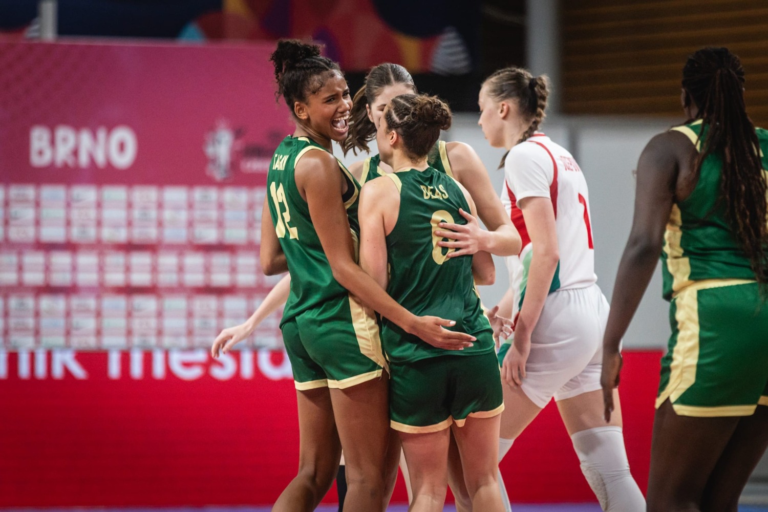 Sitaya Fagan in green, her name and number visible on her back, laughing as she embraces two Australian team-mates during a break in play. A Hungary player in white walks past and the arena backdrop reads BRNO.