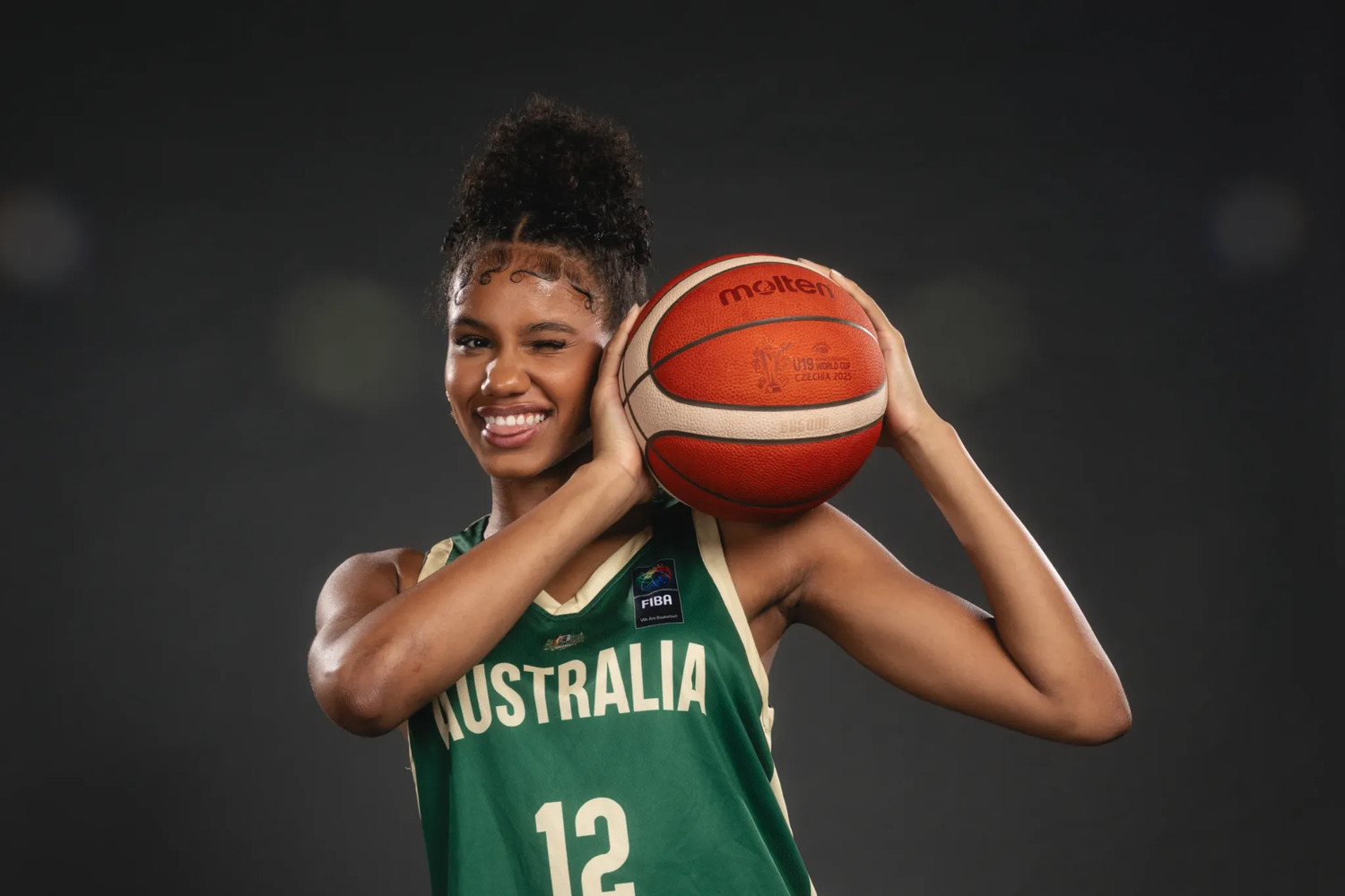 Sitaya Fagan in a green Australia number 12 jersey, smiling and holding a Molten basketball against her right shoulder with both hands, against a dark studio background. The ball is printed U19 World Cup Czechia 2025.