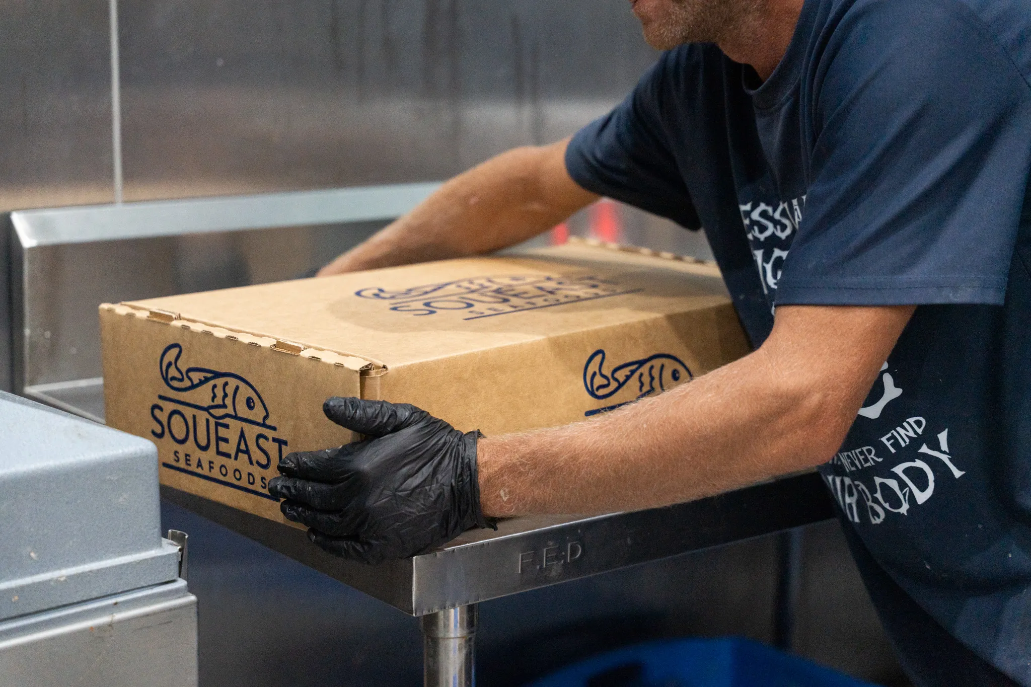 Person wearing black gloves handling a large cardboard box labeled 'Soueast Seafoods' on a metal table.