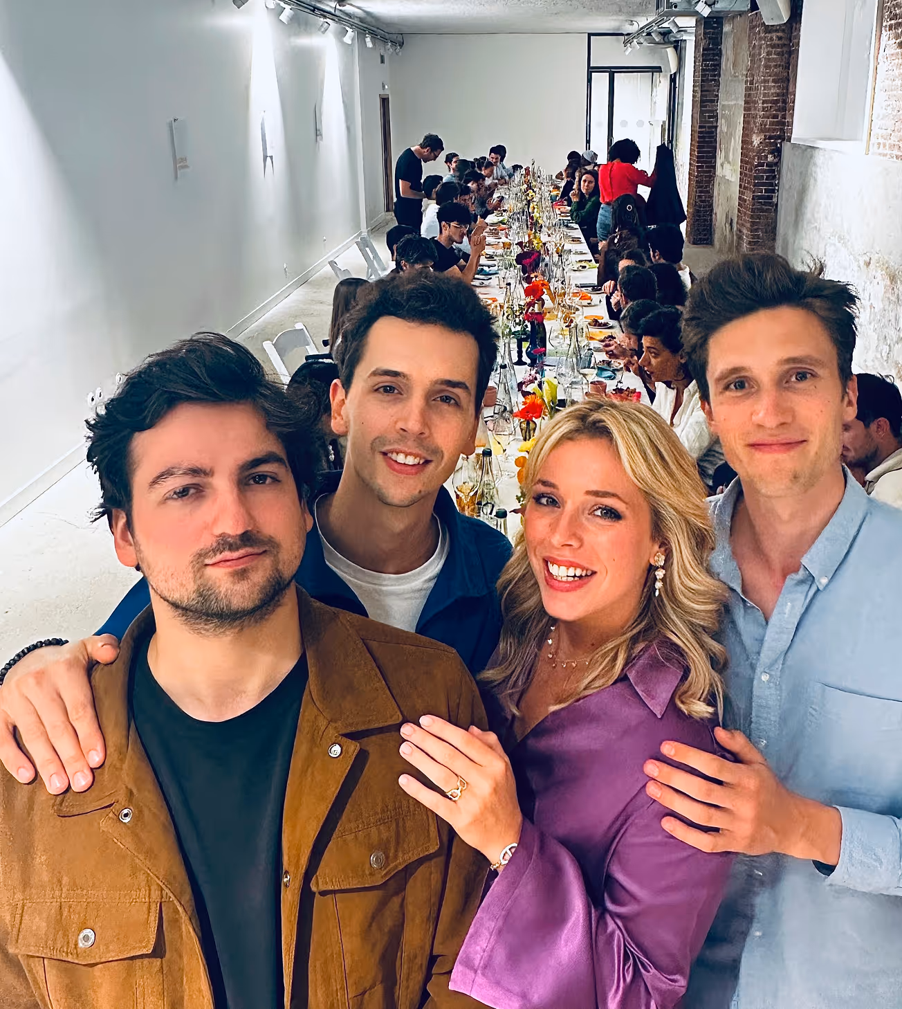 Quatre amis souriants posant devant une longue table de repas décorée dans une salle lumineuse avec murs en briques.