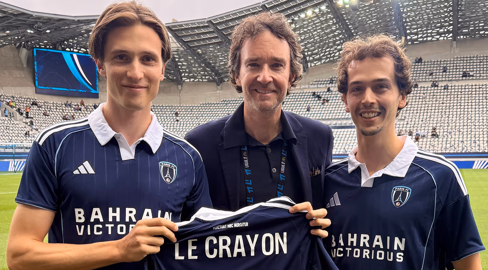 Trois hommes souriants dans un stade, deux portant des maillots de football Paris FC tenant un maillot avec l'inscription 'LE CRAYON'.