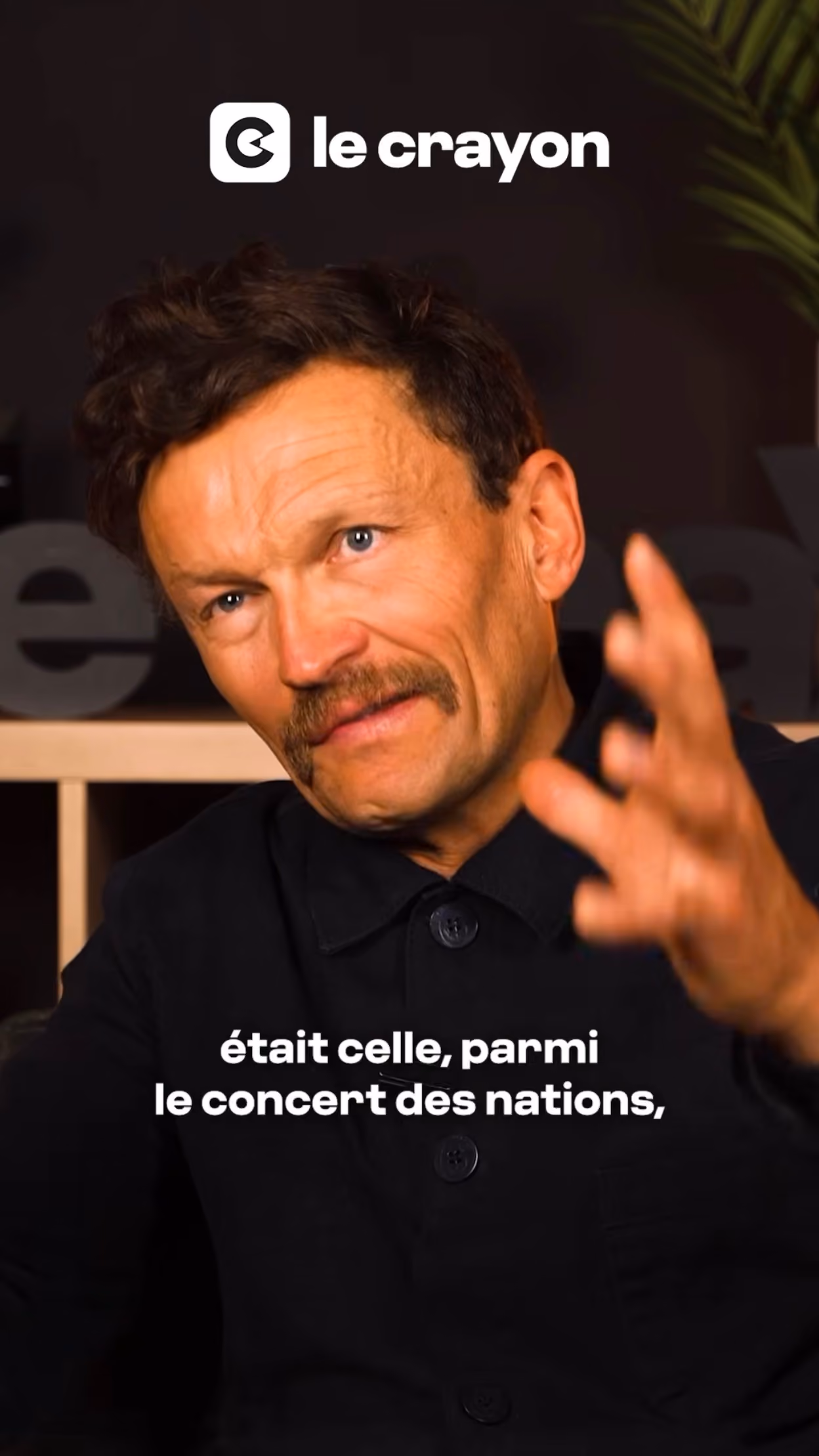 Homme moustachu en chemise noire parlant et gesticulant devant un fond sombre avec le logo Le Crayon.