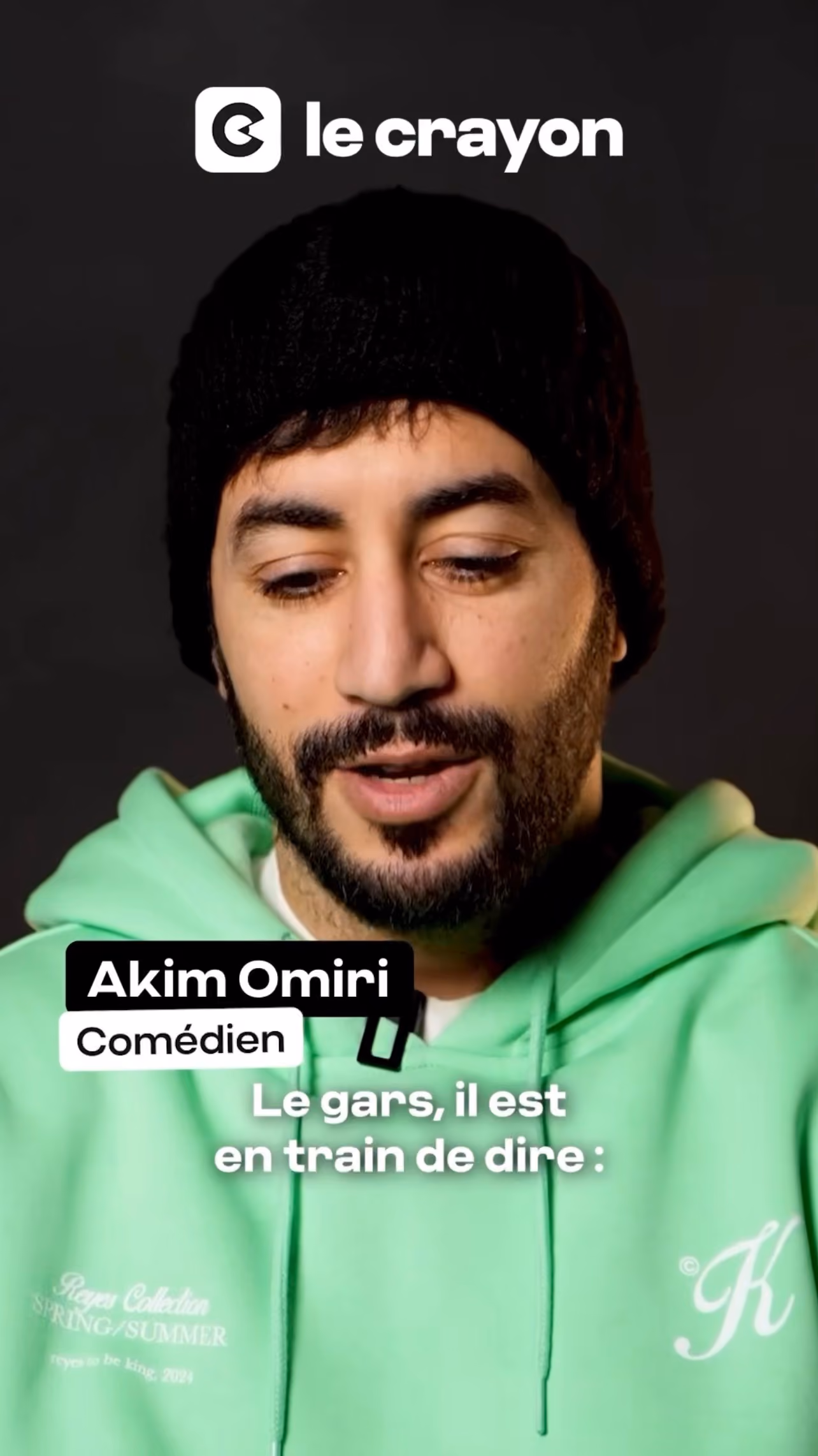 Portrait d'Akim Omiri portant un bonnet noir et un sweat à capuche vert clair, regardant vers le bas.
