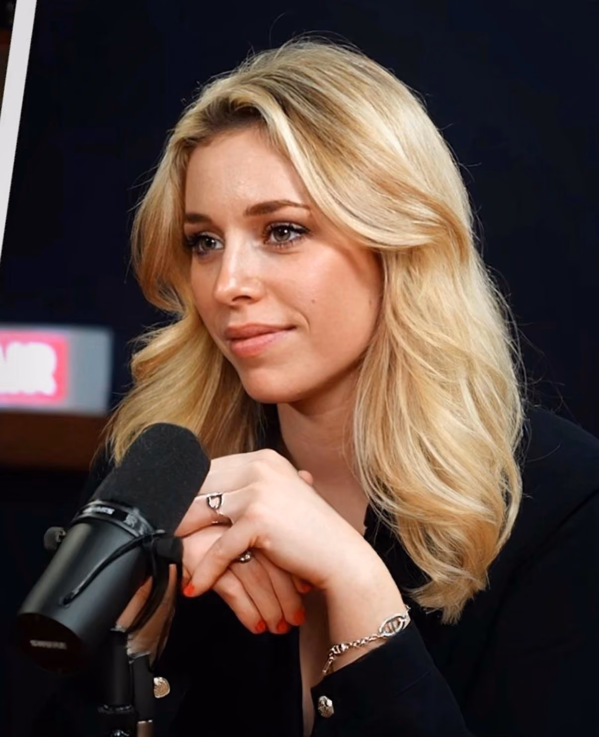 Femme blonde parlant dans un micro avec un bracelet et une bague, fond sombre.