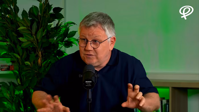 Homme avec lunettes parlant devant un micro dans une pièce avec éclairage vert et plantes.
