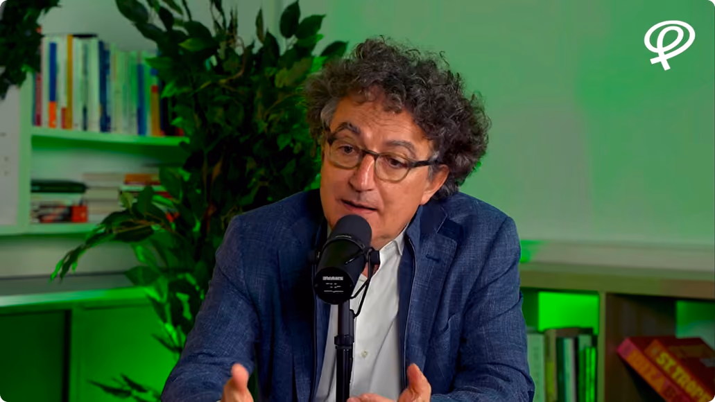 Un homme aux cheveux bouclés et lunettes parle devant un micro avec une étagère verte en arrière-plan.