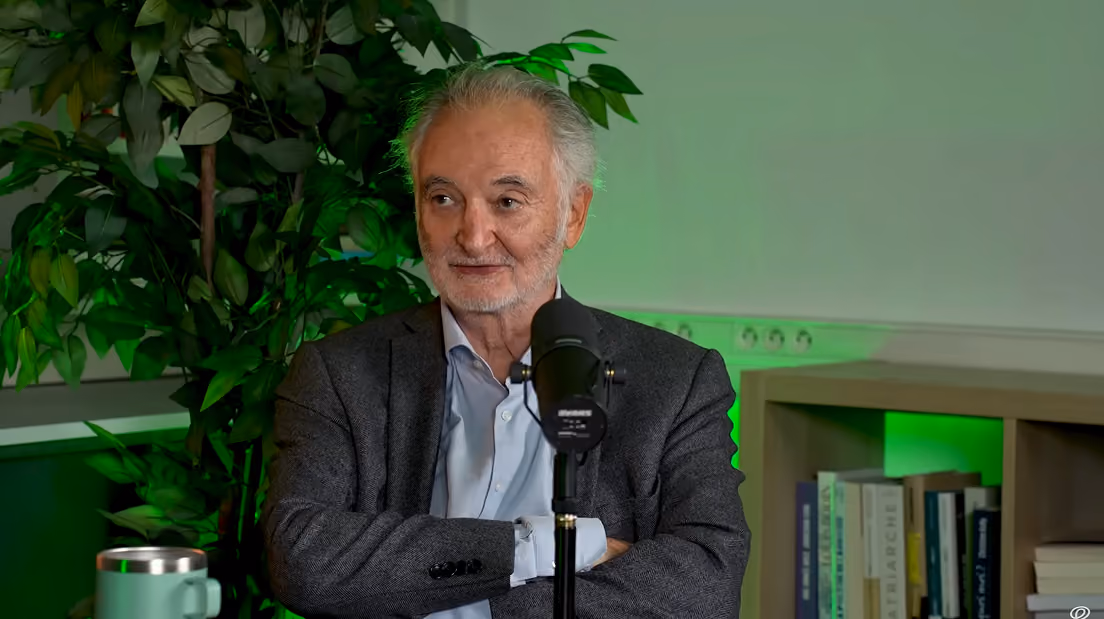 Homme âgé souriant assis devant un micro avec un mur vert, plante et étagère de livres en arrière-plan.