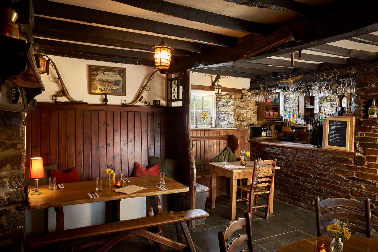 Cozy rustic pub interior with stone bar, wooden stools, hanging mugs, and a bartender behind the counter.