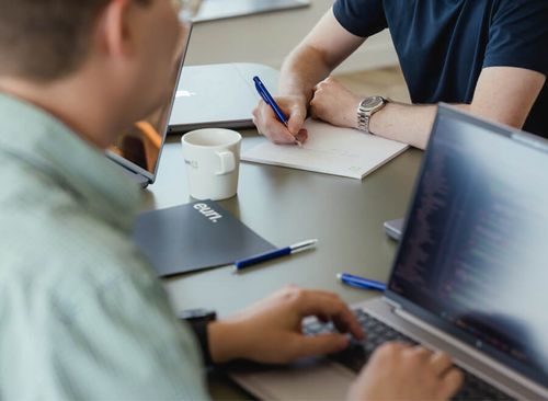 Person writing while the other person is working on their laptop.