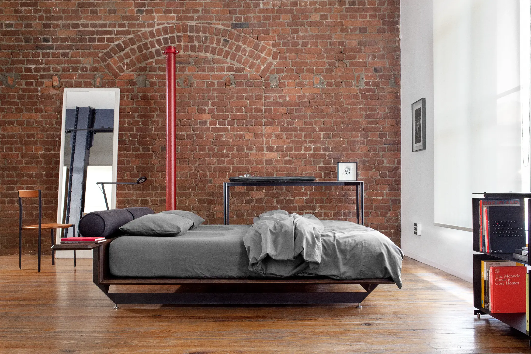 Modern bedroom with wooden floor, exposed brick wall, low bed with grey bedding, and minimalist furniture.
