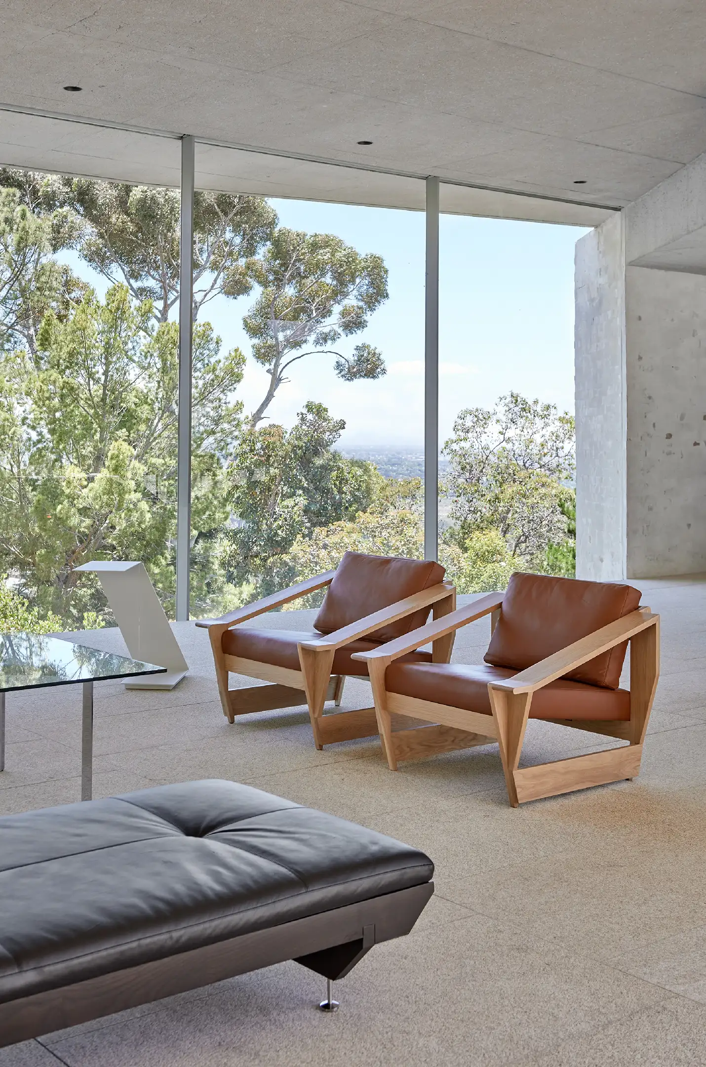 Modern lounge with two brown leather wooden armchairs and a black leather bench by floor-to-ceiling windows.