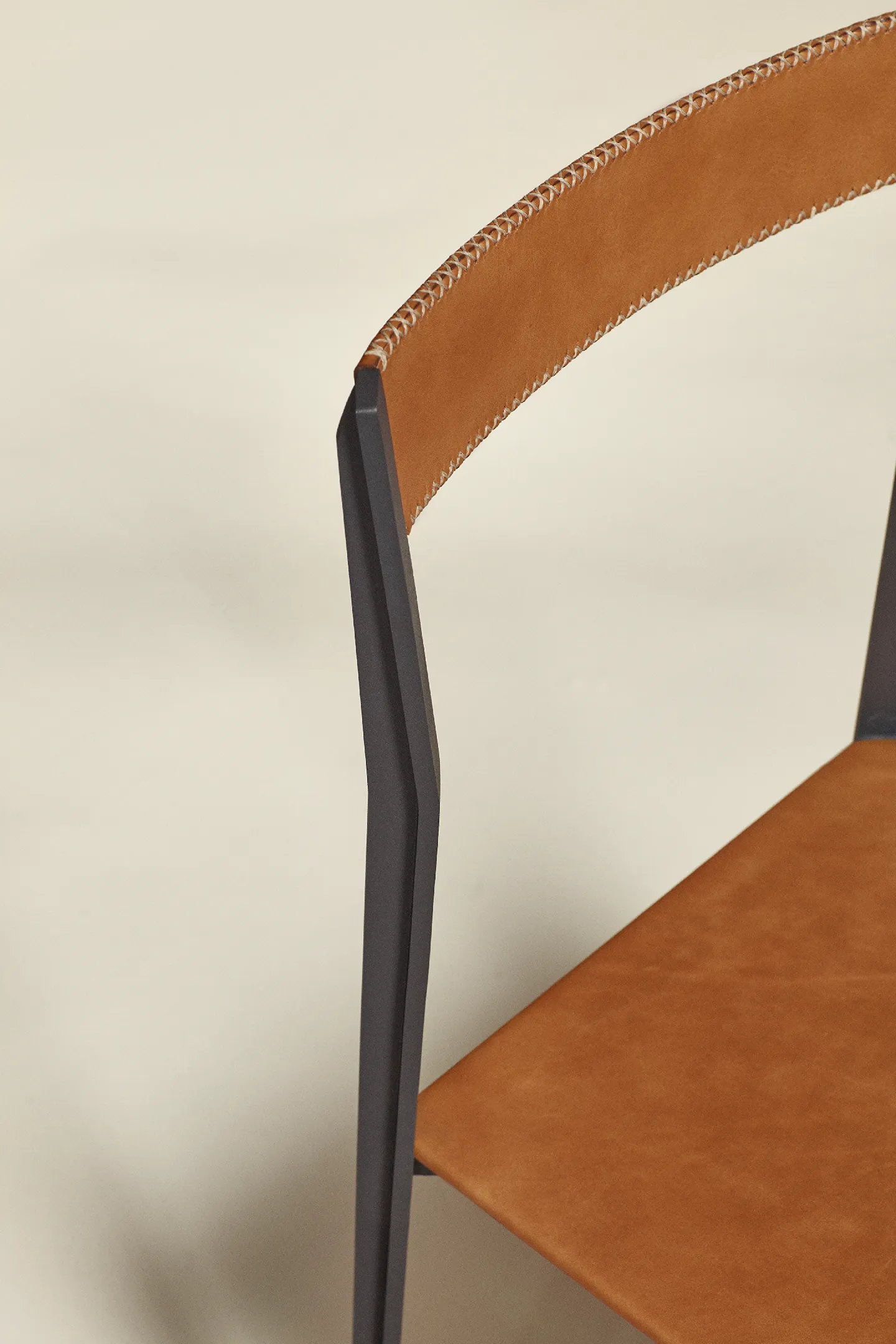 Close-up of a chair with tan leather seat and backrest with white stitching and black frame.
