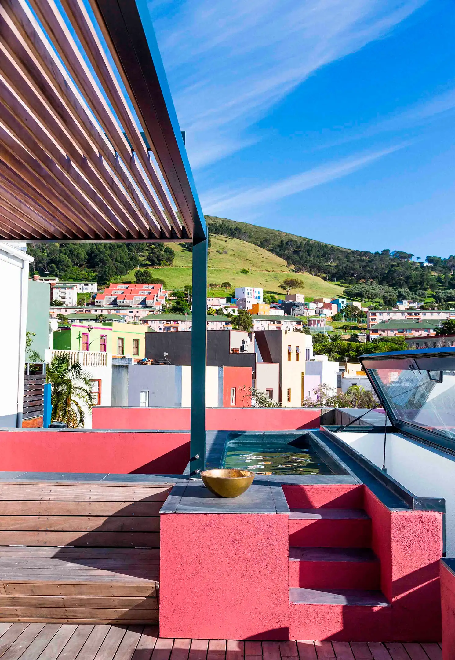 Outdoor terrace with wooden bench, red walls, water feature, and hillside homes under blue sky.