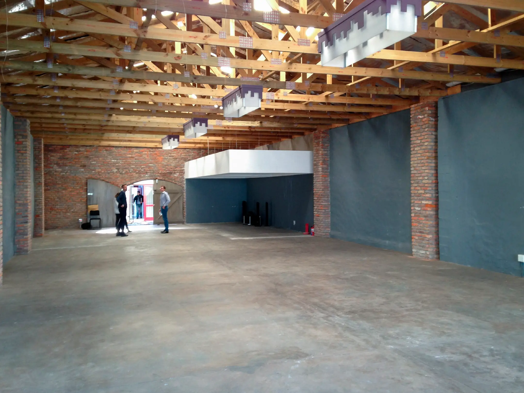 Spacious interior with exposed wooden beams, brick walls, concrete floor, and three people near the entrance.