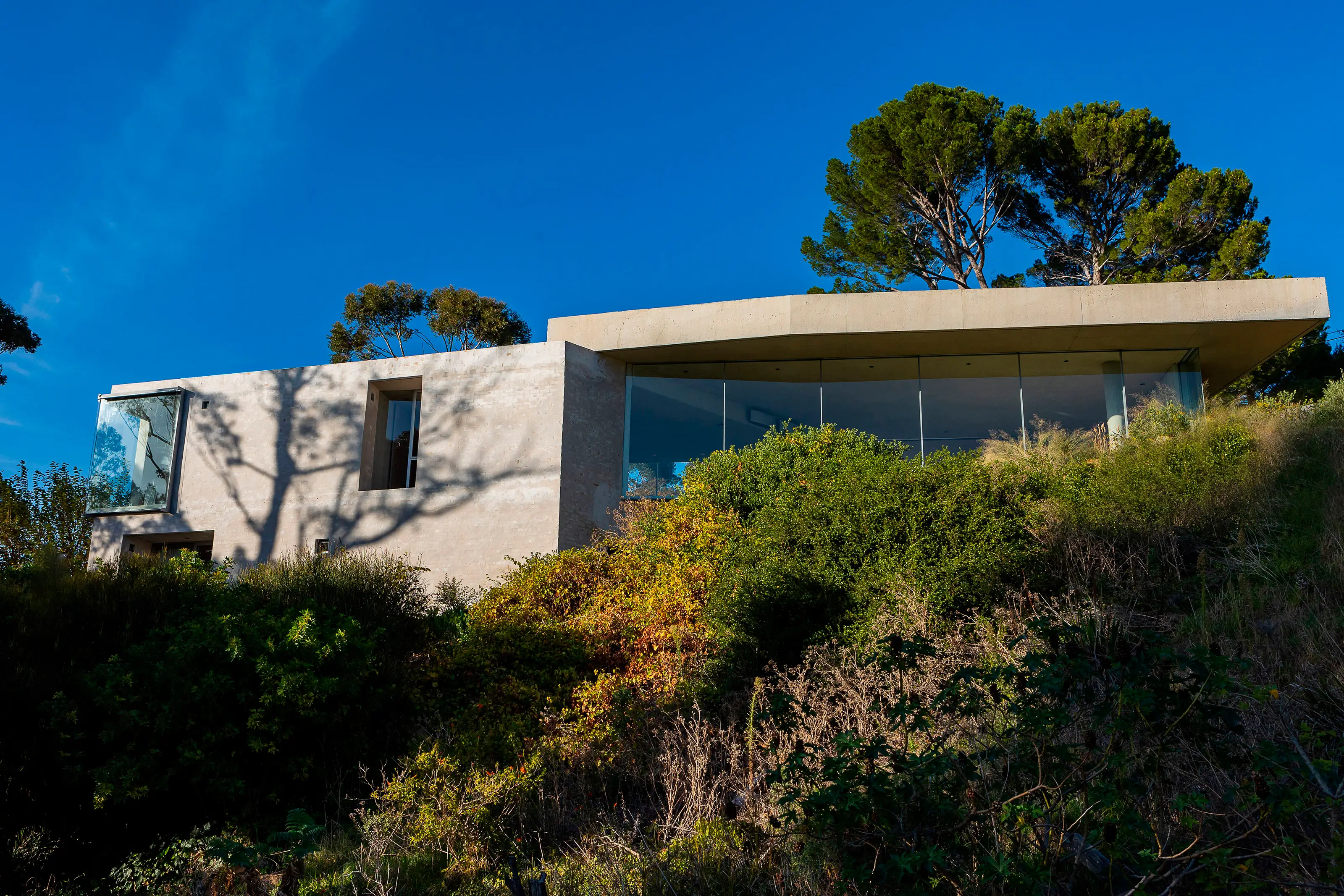 Modern concrete house with large glass windows on a hillside surrounded by green shrubs and trees.