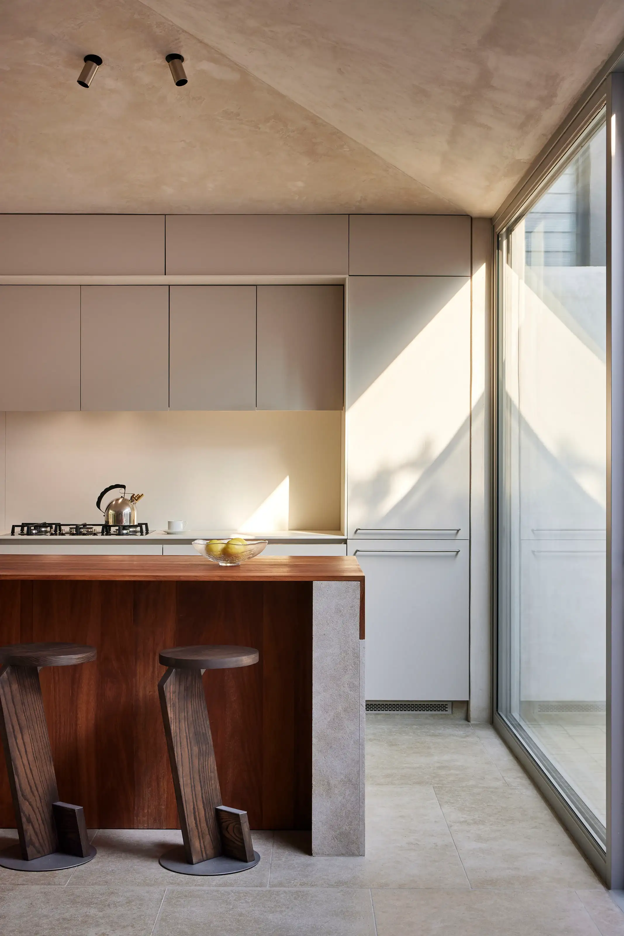 Minimalist kitchen with wooden island, two modern stools, metal kettle, and large glass window.