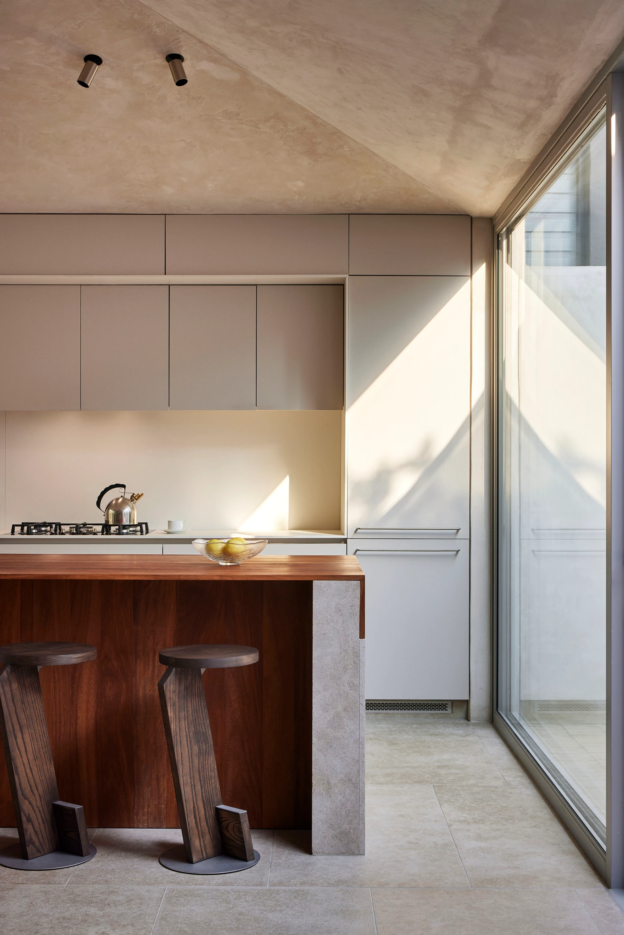 Minimalist kitchen with wooden island, two modern stools, metal kettle, and large glass window.