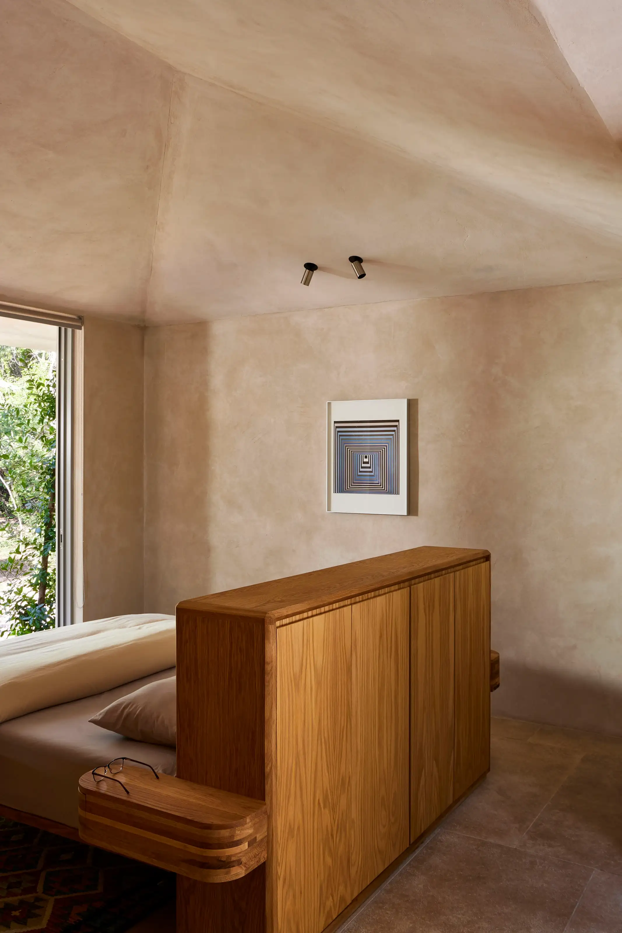 Minimalist bedroom with wooden bed frame, beige walls, window showing greenery, and geometric wall art.