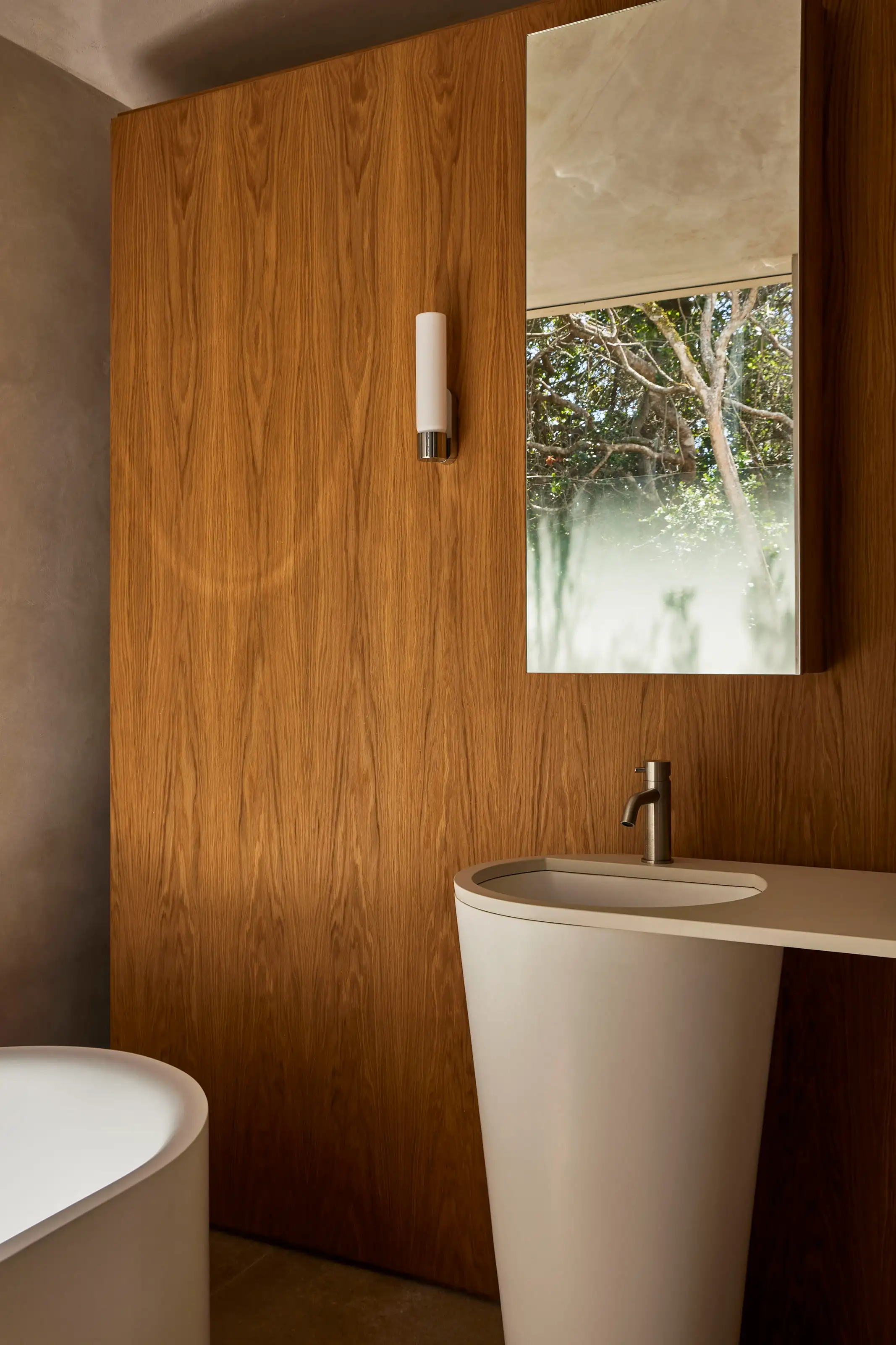 Modern bathroom with wooden wall, white freestanding sink, bathtub, and mirror reflecting trees outside.
