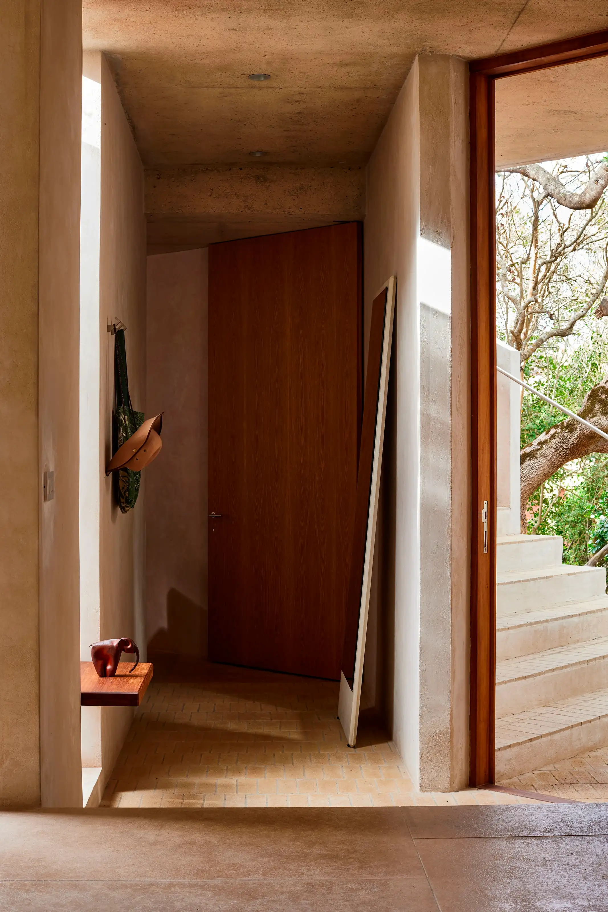 Minimalist hallway with wooden door, leaning mirror, hat and bag on wall hooks, and outdoor stairs visible.