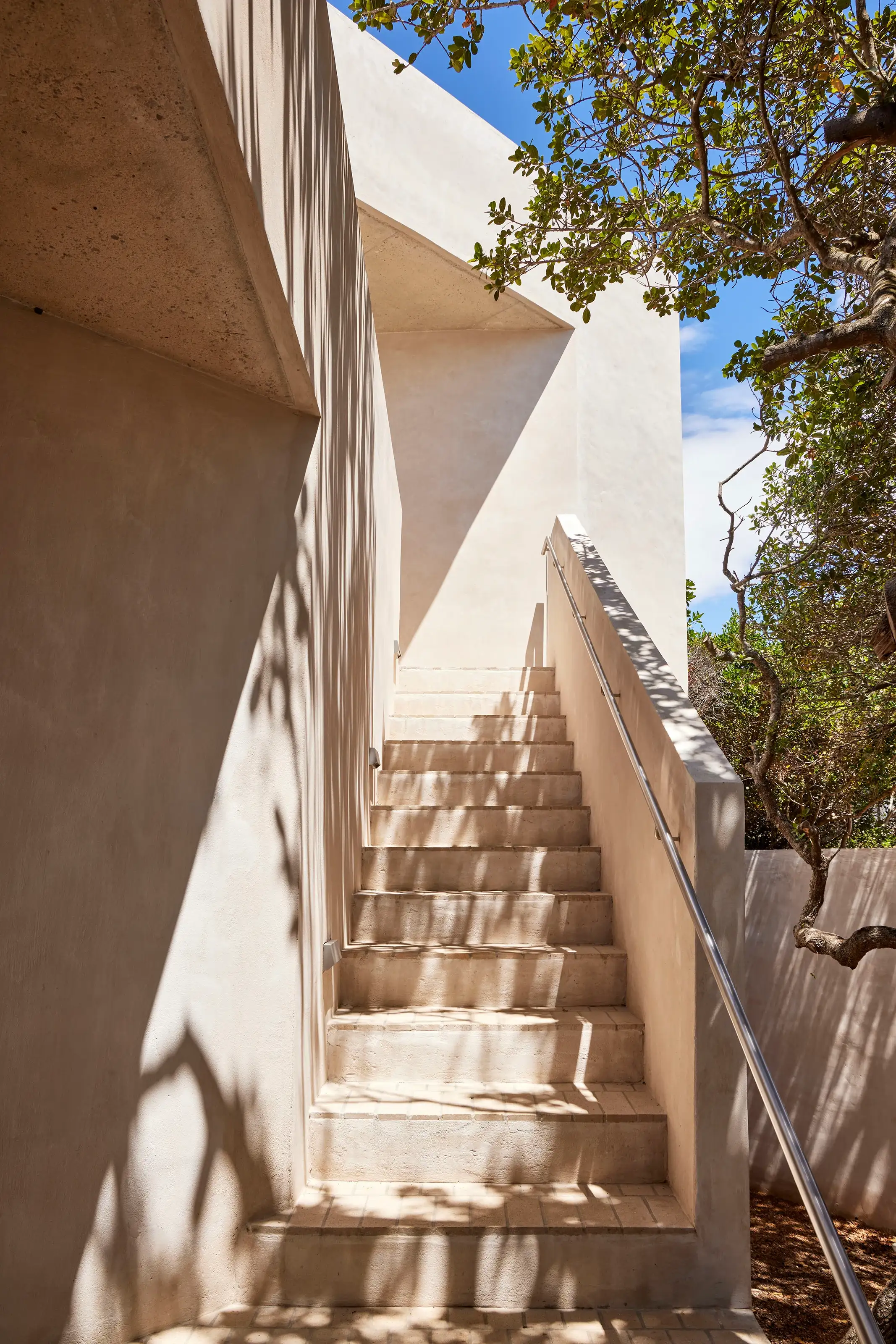 Sunlit outdoor stairs with a metal handrail and tree shadows on the beige walls and steps.