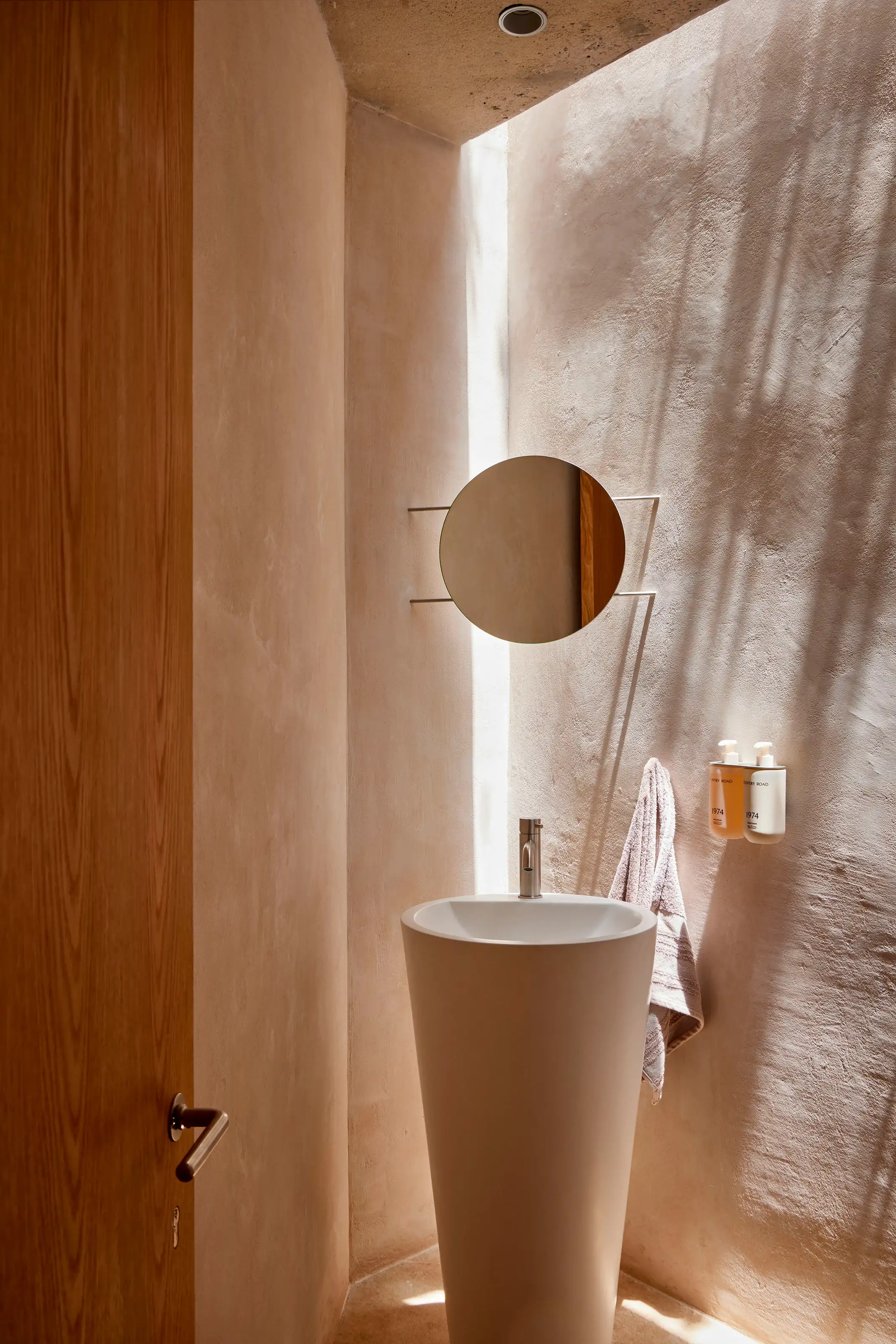 Minimalist bathroom with tall white pedestal sink, round mirror, towel, and soap dispensers on textured wall.