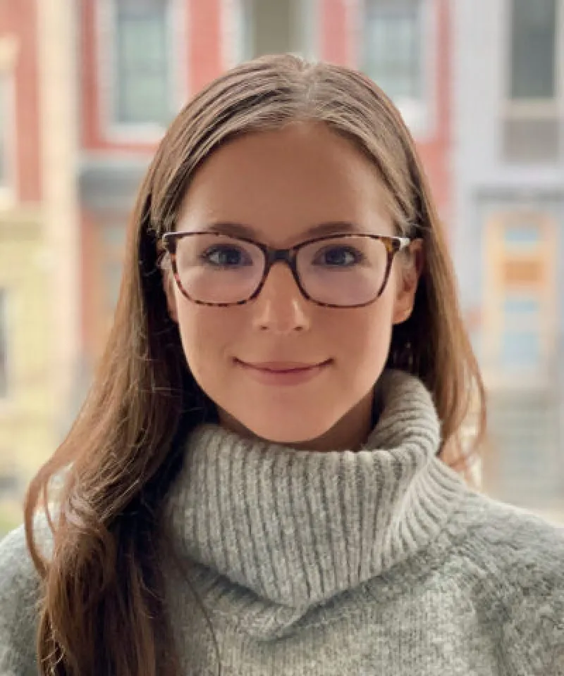 Young woman with long brown hair and glasses wearing a thick gray turtleneck sweater, smiling softly with a blurred urban background.