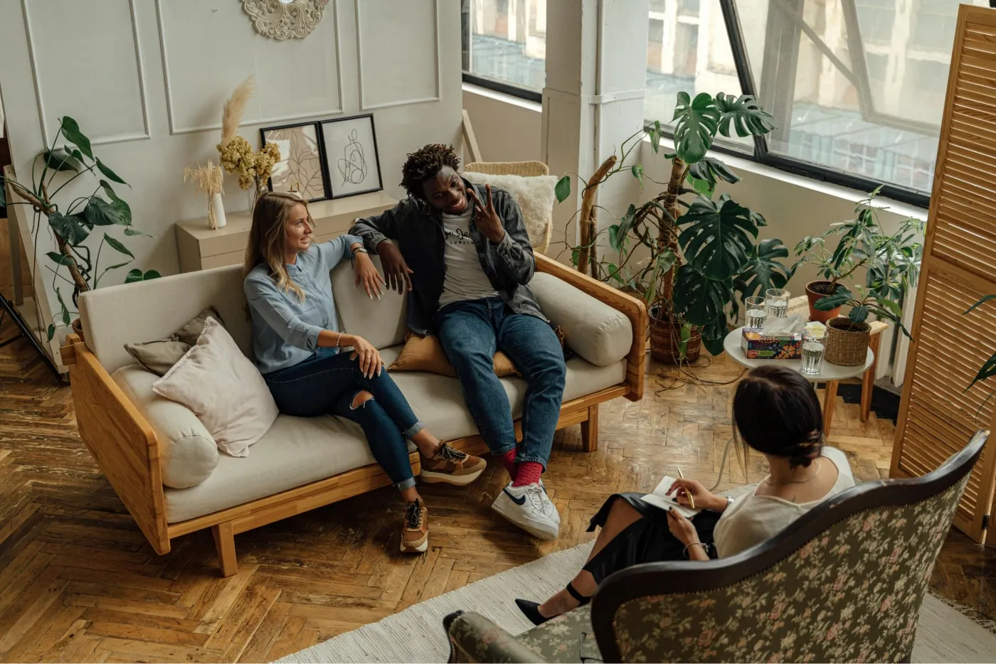 A couple sitting on a beige couch in a cozy, plant-filled room talking to a woman who is taking notes.