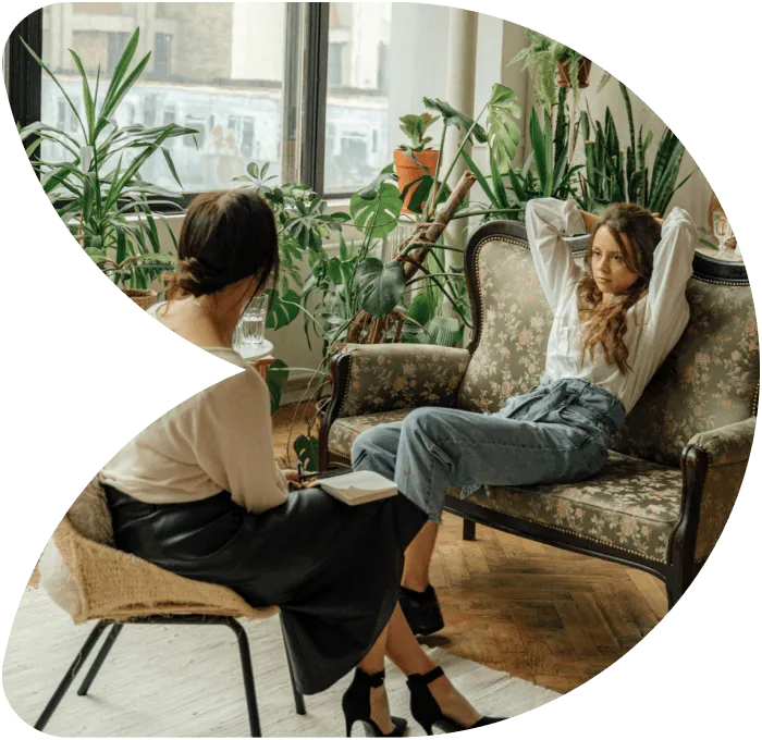 Two women in a cozy living room with many plants; one woman sits on a chair with a notebook, the other reclines on a floral sofa.