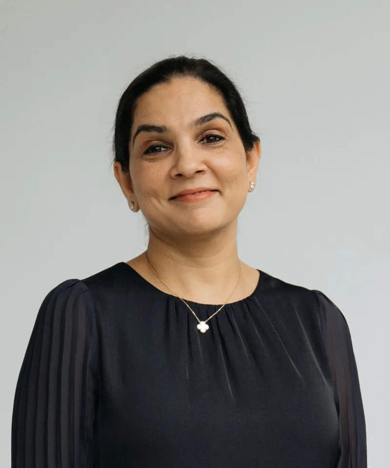 Smiling woman with dark hair pulled back, wearing a black blouse and a gold necklace with a flower-shaped pendant.