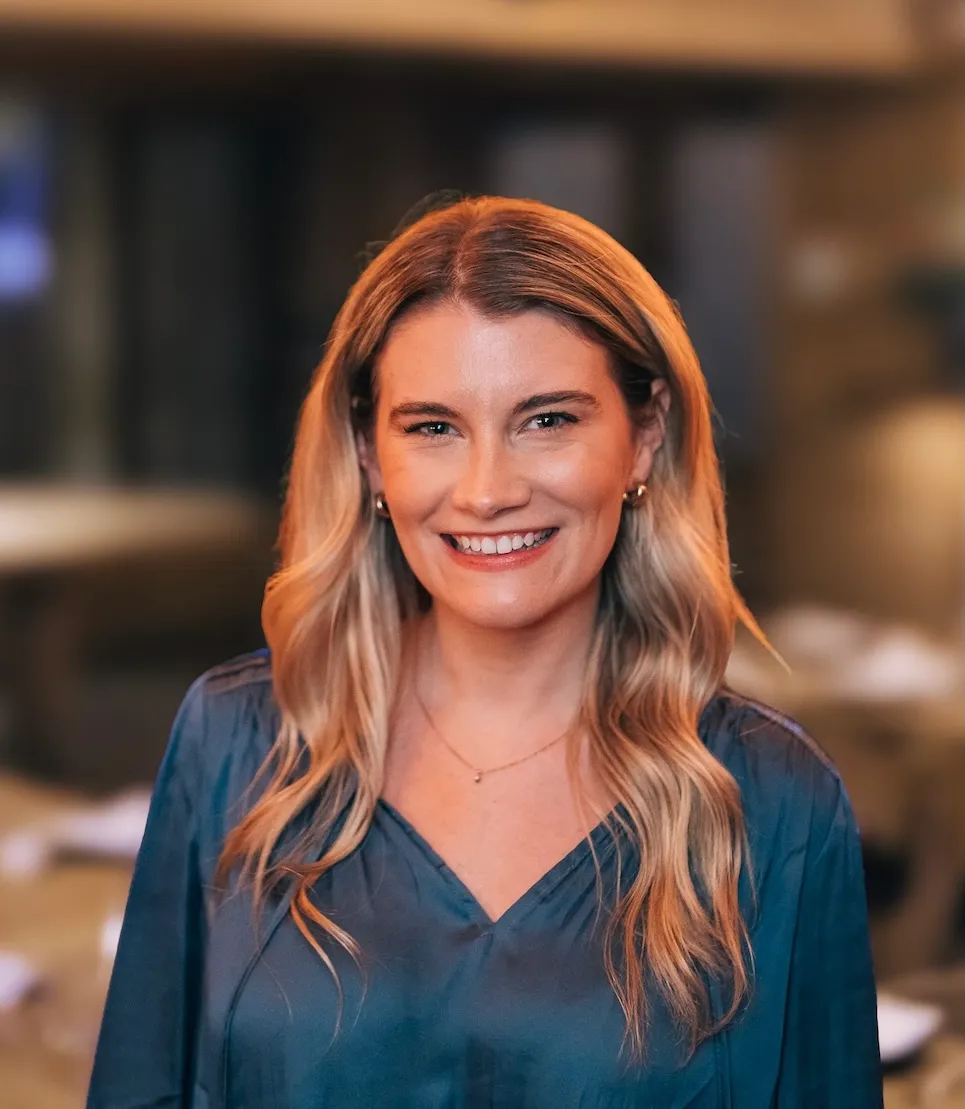 Smiling woman with long blonde hair wearing a blue blouse in a softly lit indoor setting.
