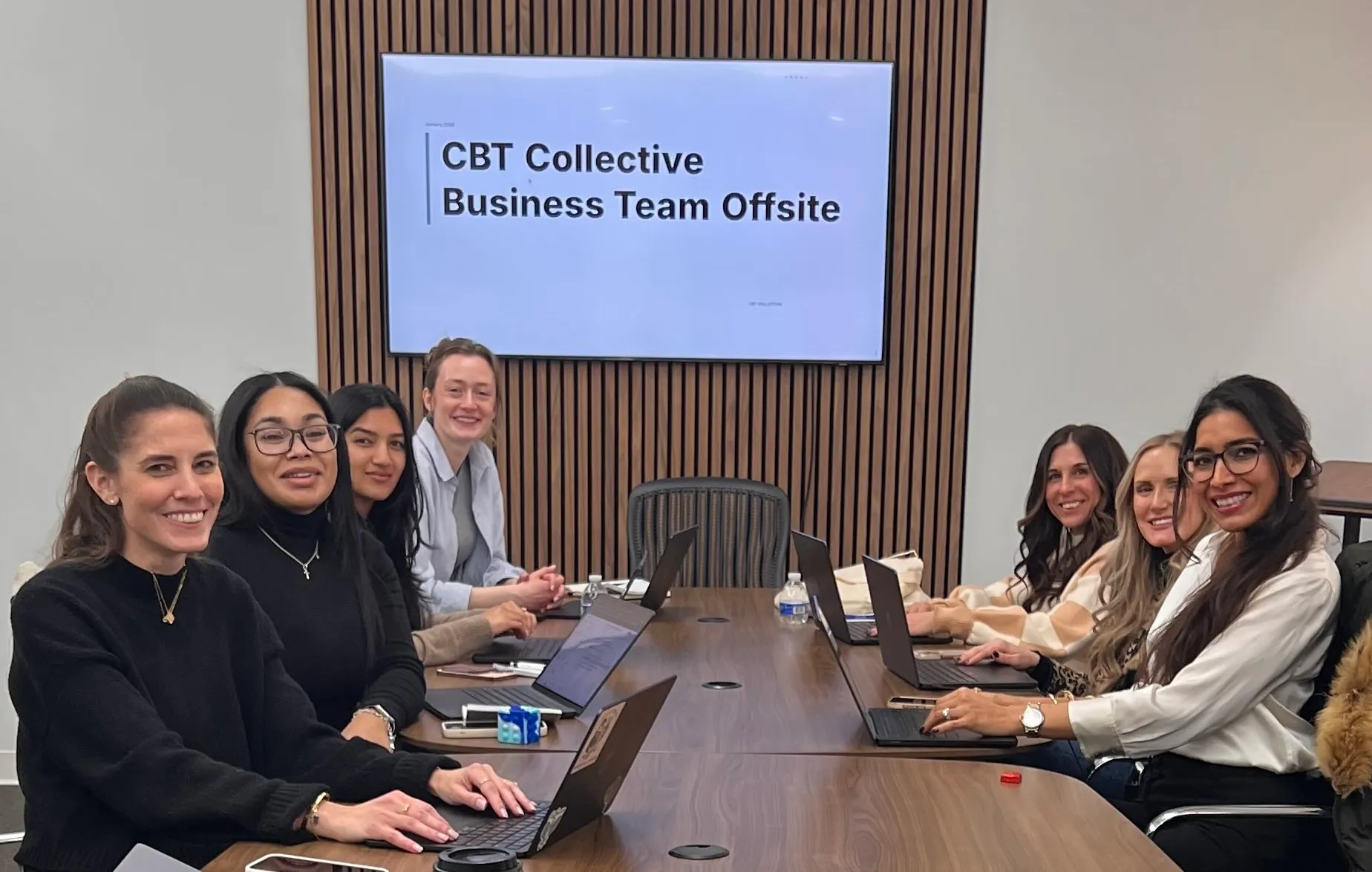 Seven women sitting at a conference table with laptops, smiling towards the camera; a screen behind them reads 'CBT Collective Business Team Offsite'.