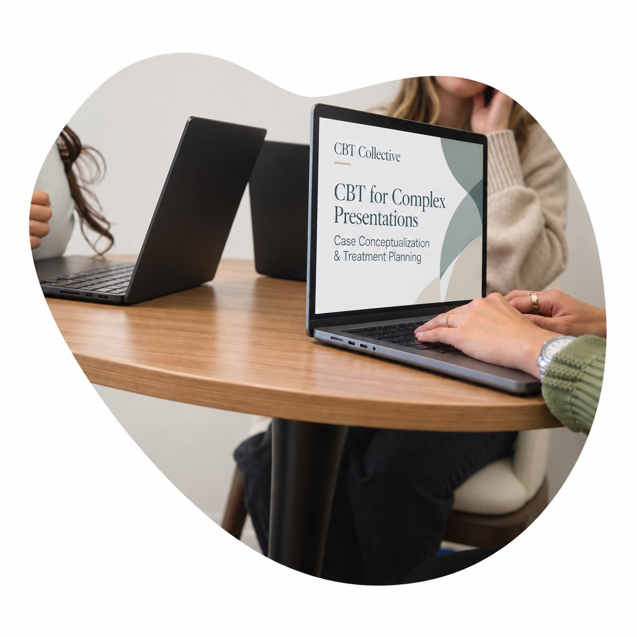 Two people sitting at a round wooden table with laptops open, one showing a screen titled 'CBT Collective: CBT for Complex Presentations, Case Conceptualization & Treatment Planning.'