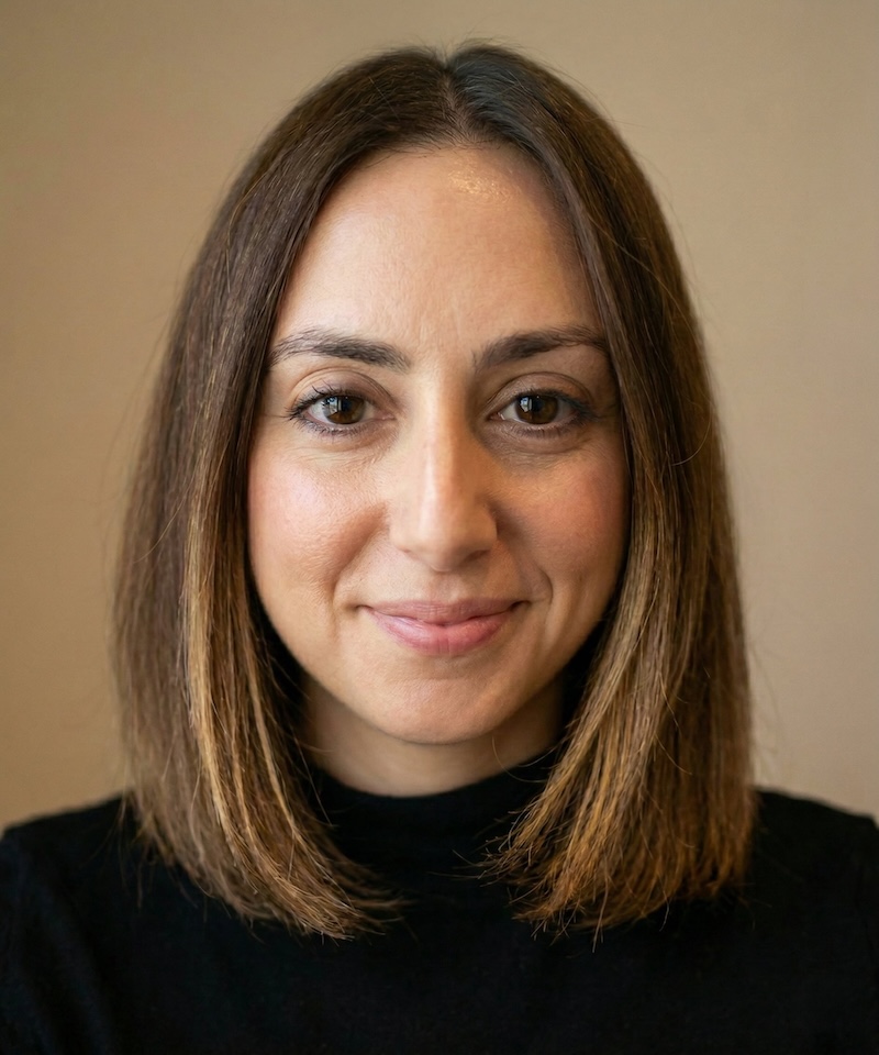 A woman with shoulder-length brown hair smiles softly at the camera. She wears a black top against a beige background, conveying a warm and friendly tone.