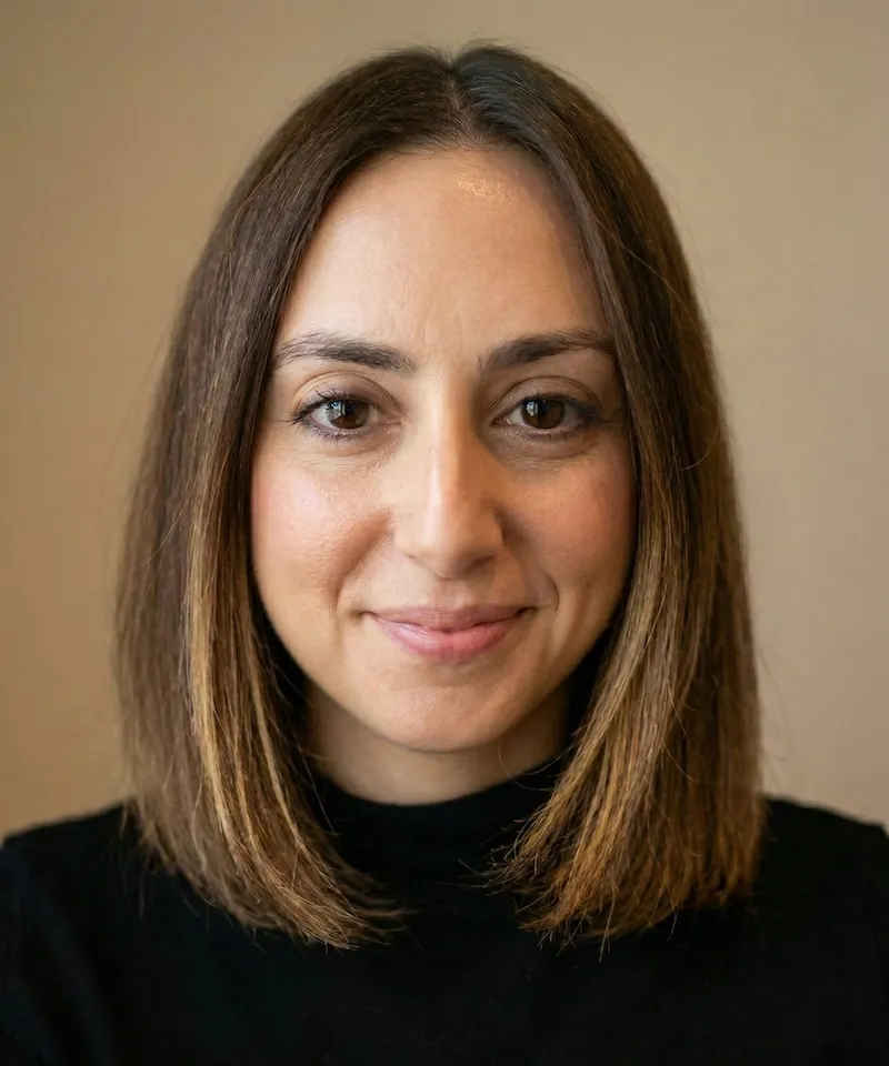 A woman with shoulder-length brown hair smiles softly at the camera. She wears a black top against a beige background, conveying a warm and friendly tone.