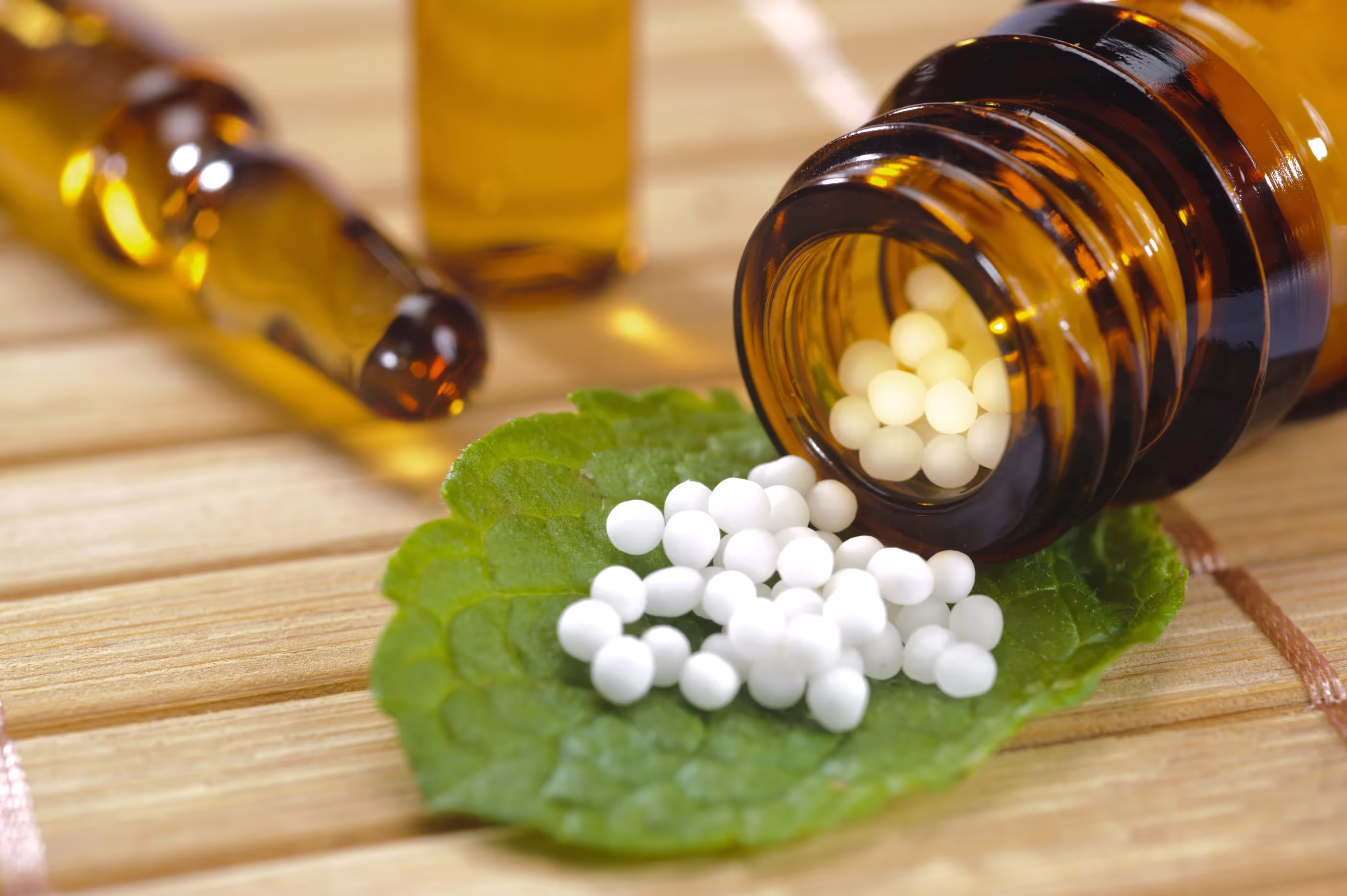 Granules homéopathiques sur une feuille de menthe, près d'un flacon et d'une ampoule en verre sur un support en bois.