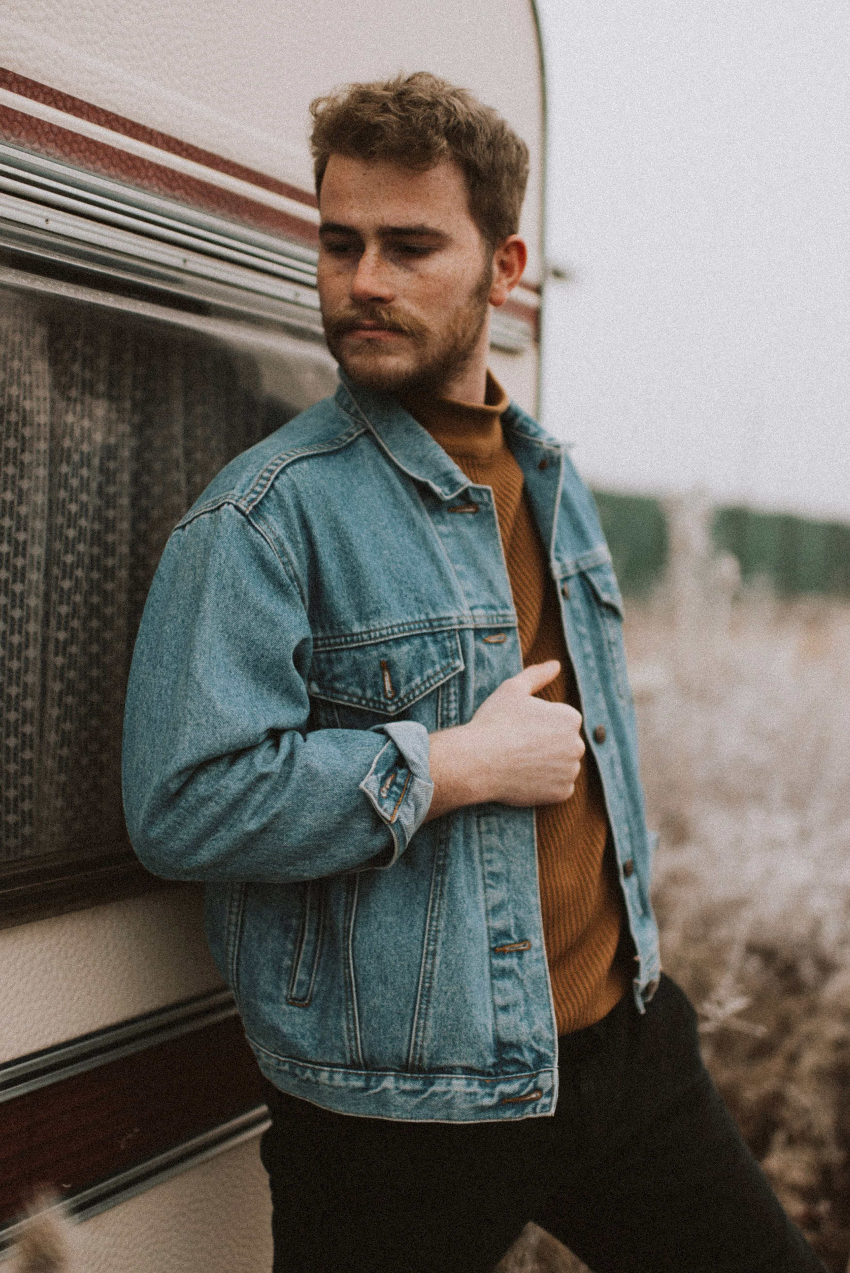Homme aux cheveux bruns bouclés et barbe soignée, look décontracté avec veste en jean et pull col roulé, en extérieur.