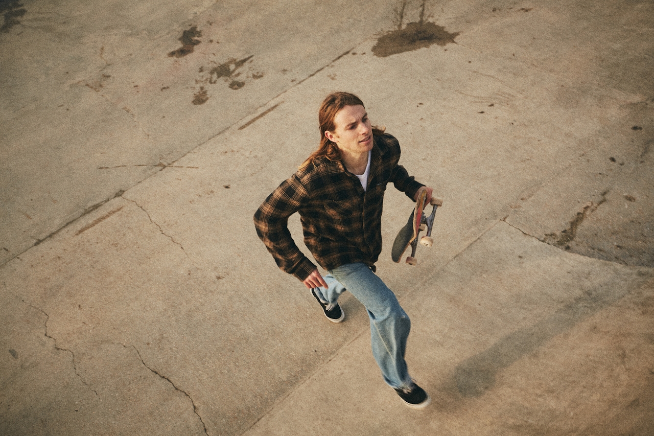 Jeune homme aux cheveux longs et denses, portant une chemise à carreaux, marchant avec un skateboard. Aspect capillaire visible.