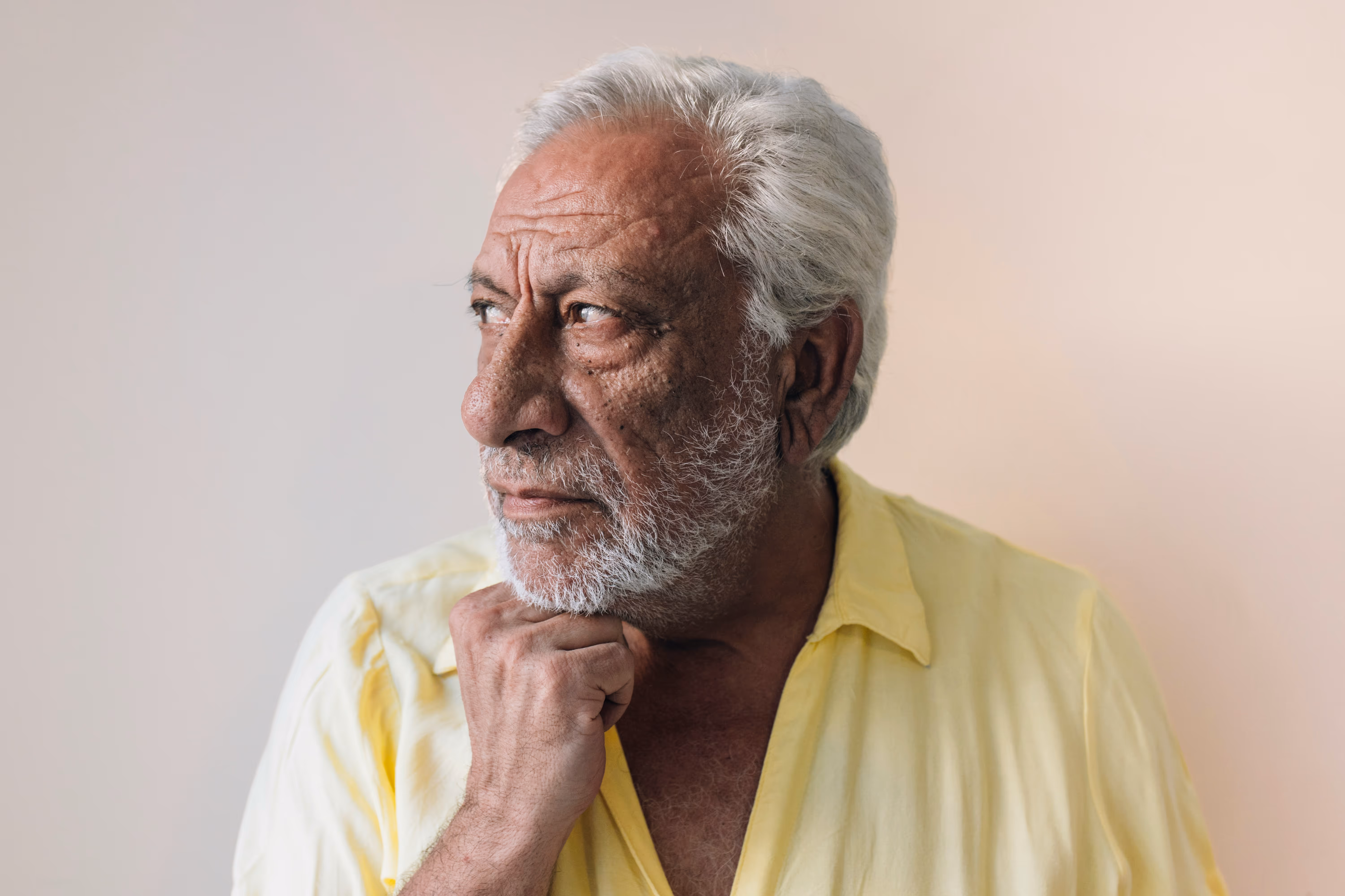 Homme âgé aux cheveux gris épais et barbe, posant la main sur son menton, contre un mur clair.