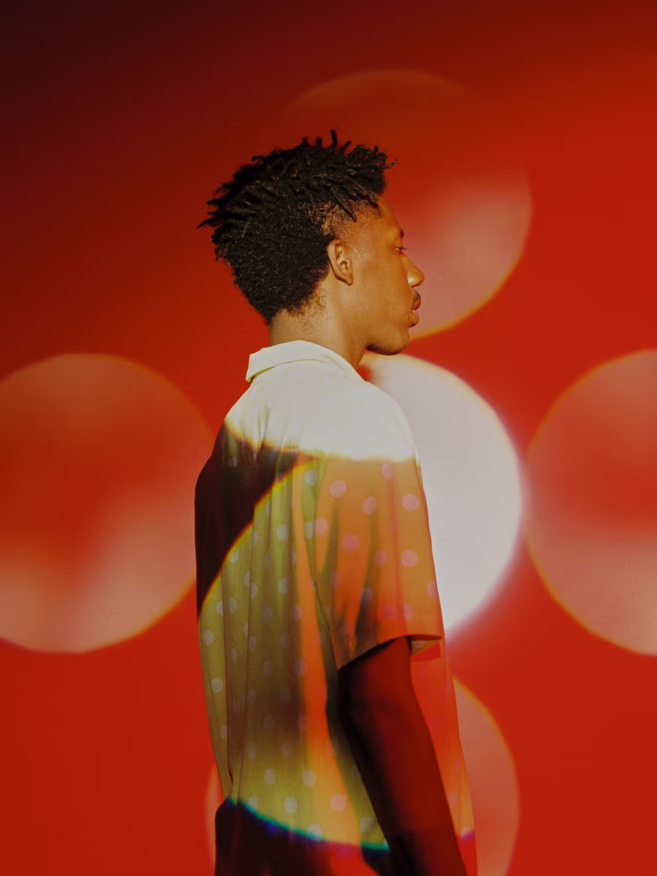 Profil d'un homme noir avec des dreadlocks et une chemise à pois, profil, fond rouge.