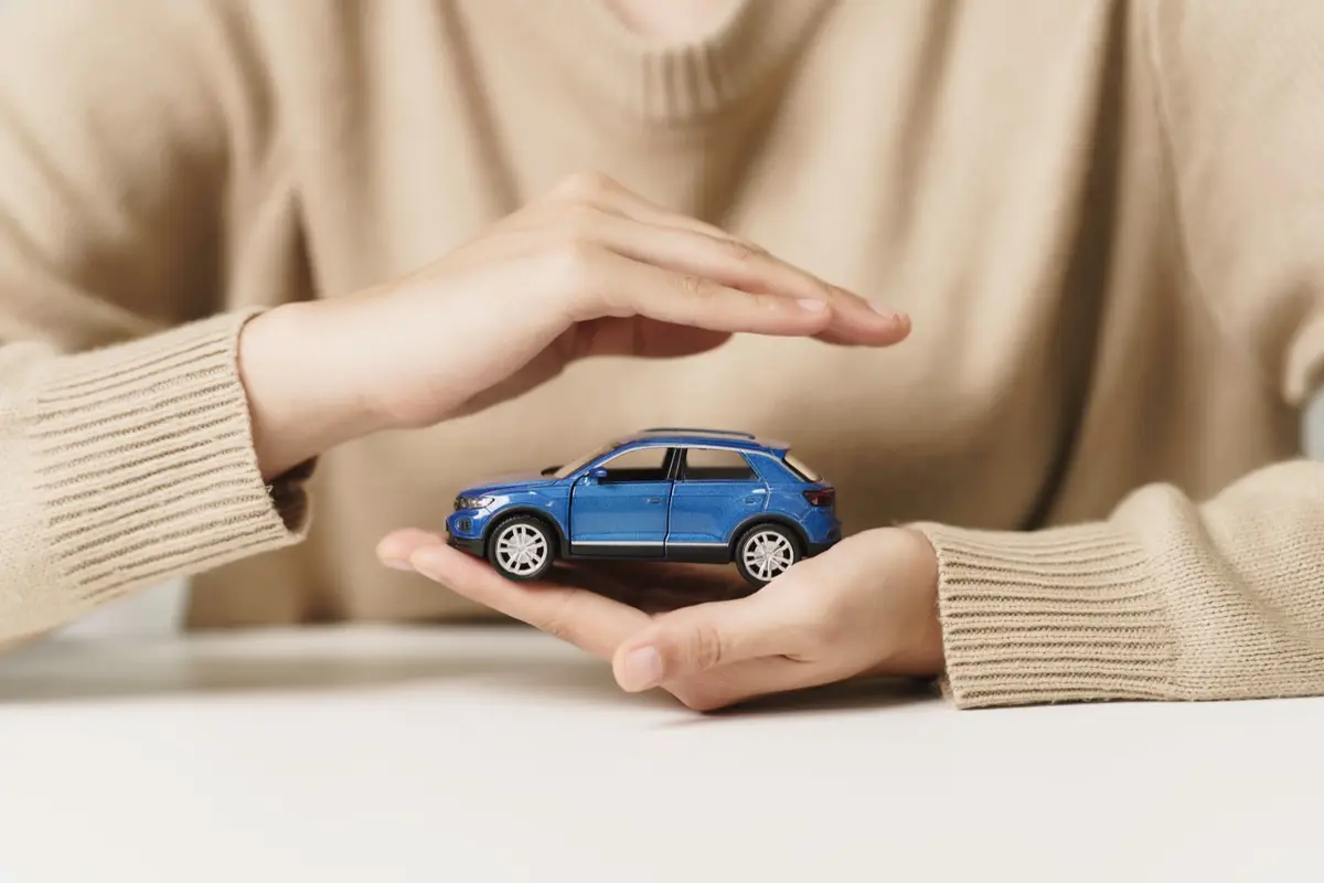 Ein blaues Auto als Figur wird schützend zwischen zwei Händen gehalten