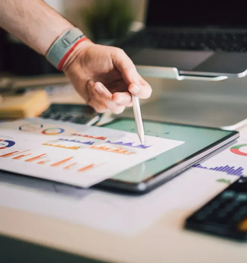A person analyzing spreadsheets on a laptop and tablet while holding printed financial bar charts at a workspace.
