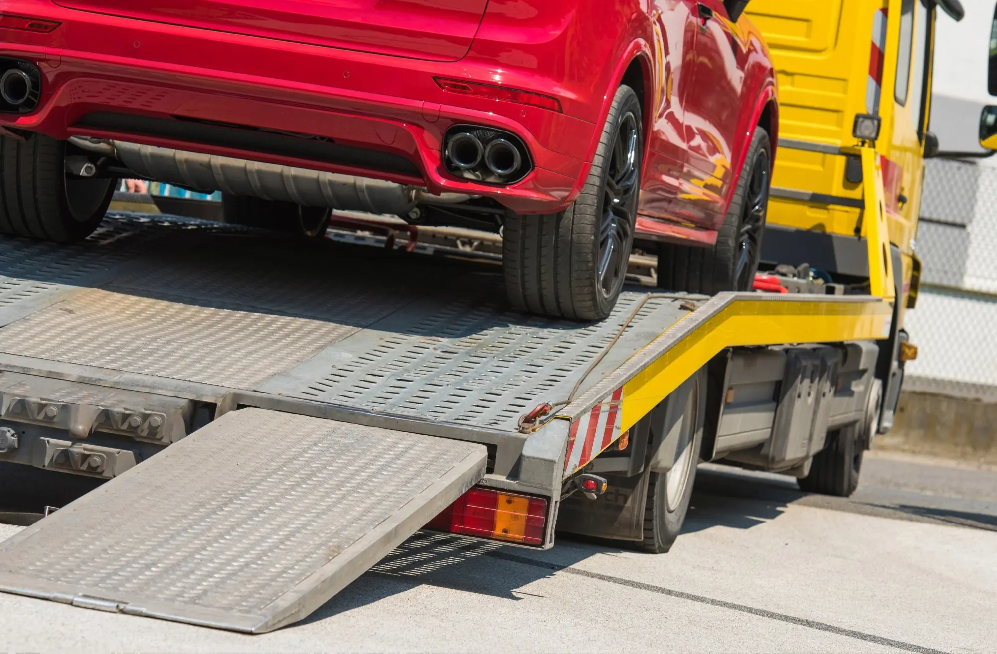 Rear of a red car being loaded onto a yellow flatbed tow truck with a metal ramp.