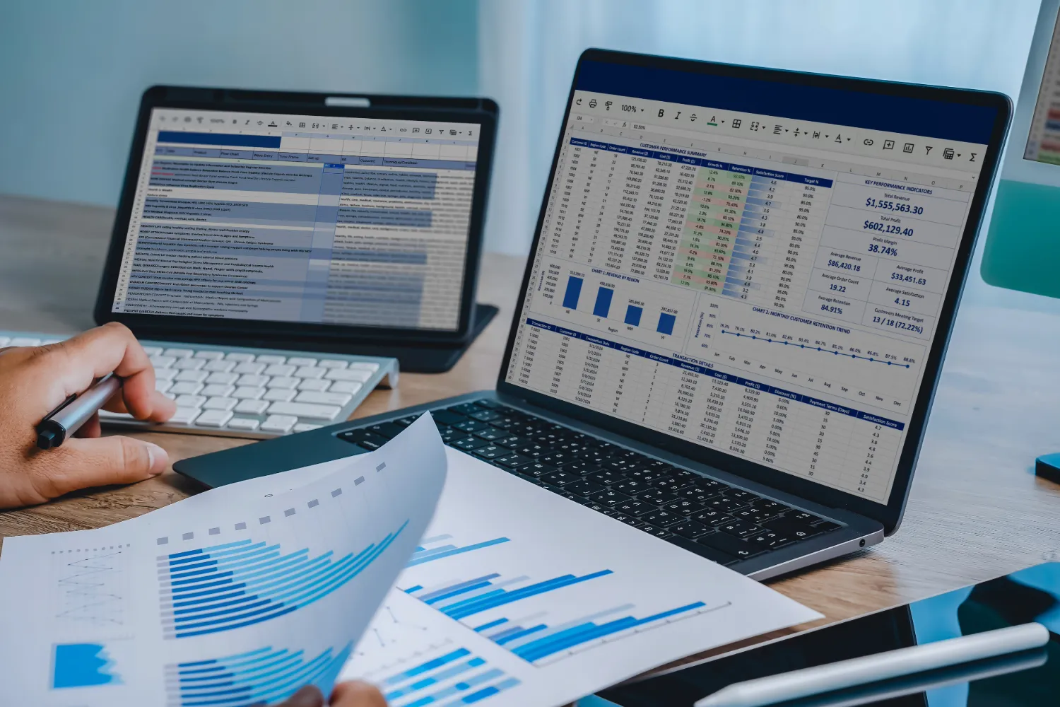 A person analyzing spreadsheets on a laptop and tablet while holding printed financial bar charts at a workspace.
