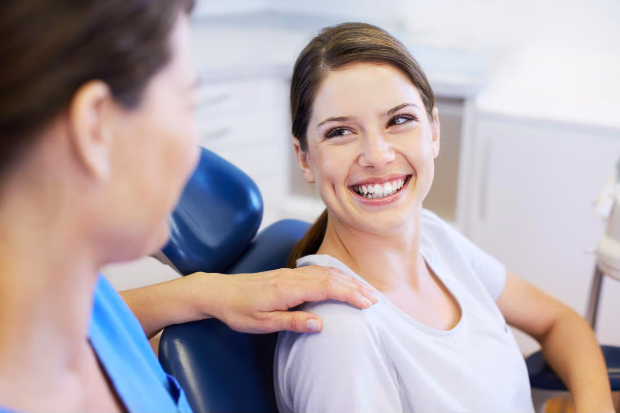 Smiling woman in a dental chair looks at a person comforting her with a hand on her shoulder.