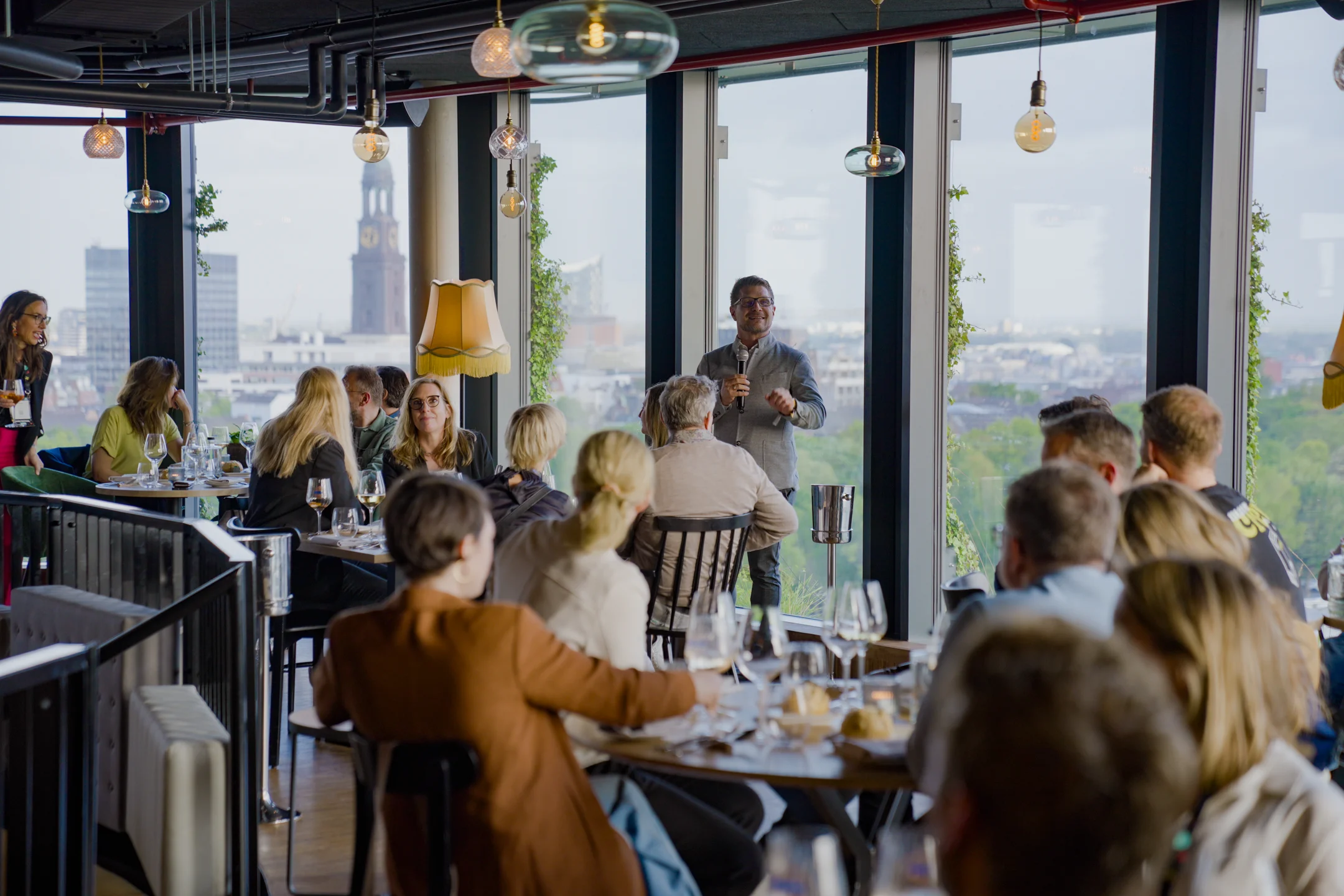 Mann hält eine Rede mit Mikrofon vor einem Publikum in einem Restaurant mit großen Fenstern und Blick auf eine Stadt.