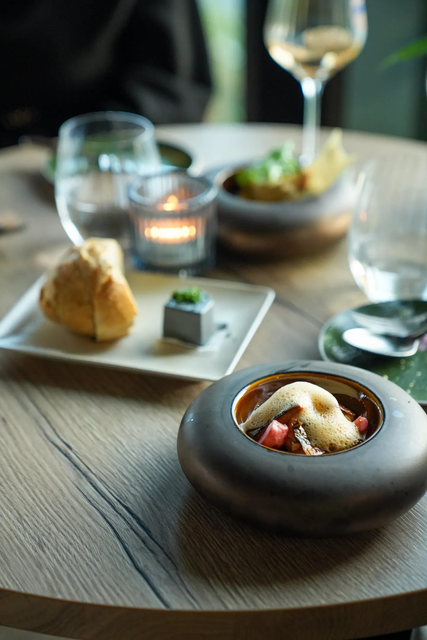 Eleganter gedeckter Tisch mit einem Gericht in einer runden, grauen Schüssel mit Schaum, Brot auf einem weißen Teller und einem Weinglas im Hintergrund.
