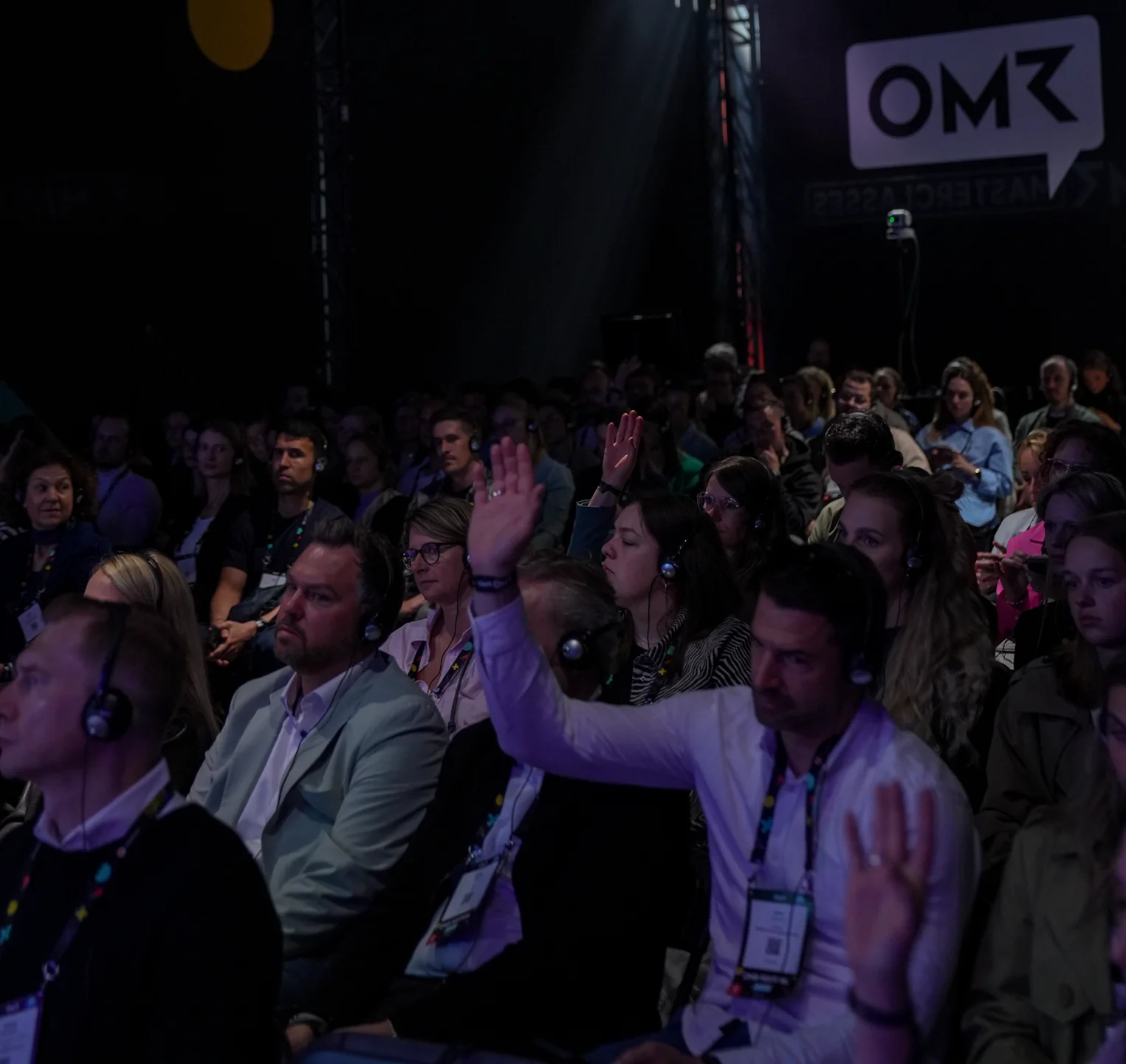 Teilnehmer bei einer Konferenz sitzen mit Kopfhörern im Publikum, einige heben die Hand, vor dunklem Hintergrund mit OMR-Logo.