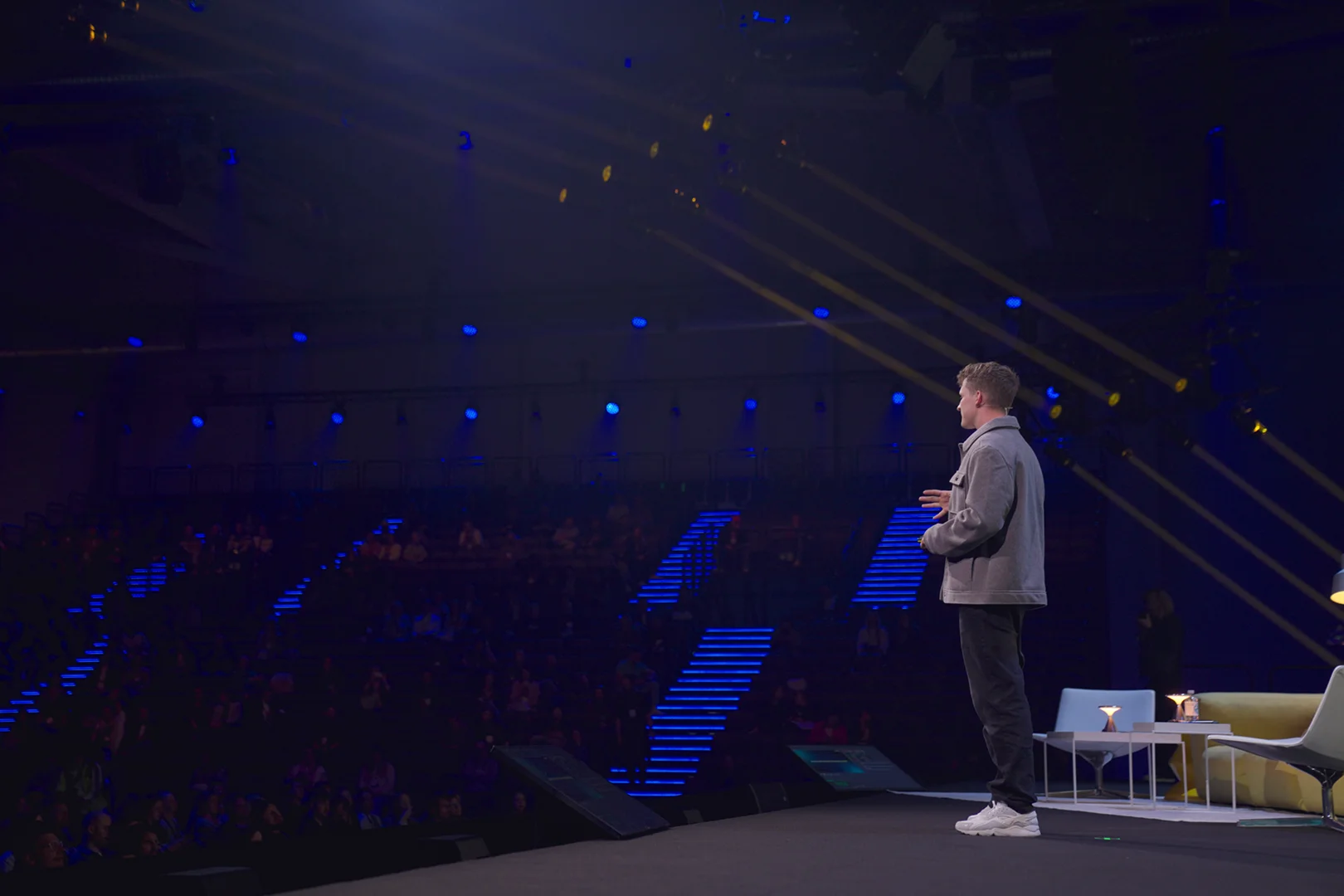 Ein junger Mann steht auf einer beleuchteten Bühne und spricht vor einem Publikum in einem großen, dunklen Saal mit blauen Treppenlichtern.